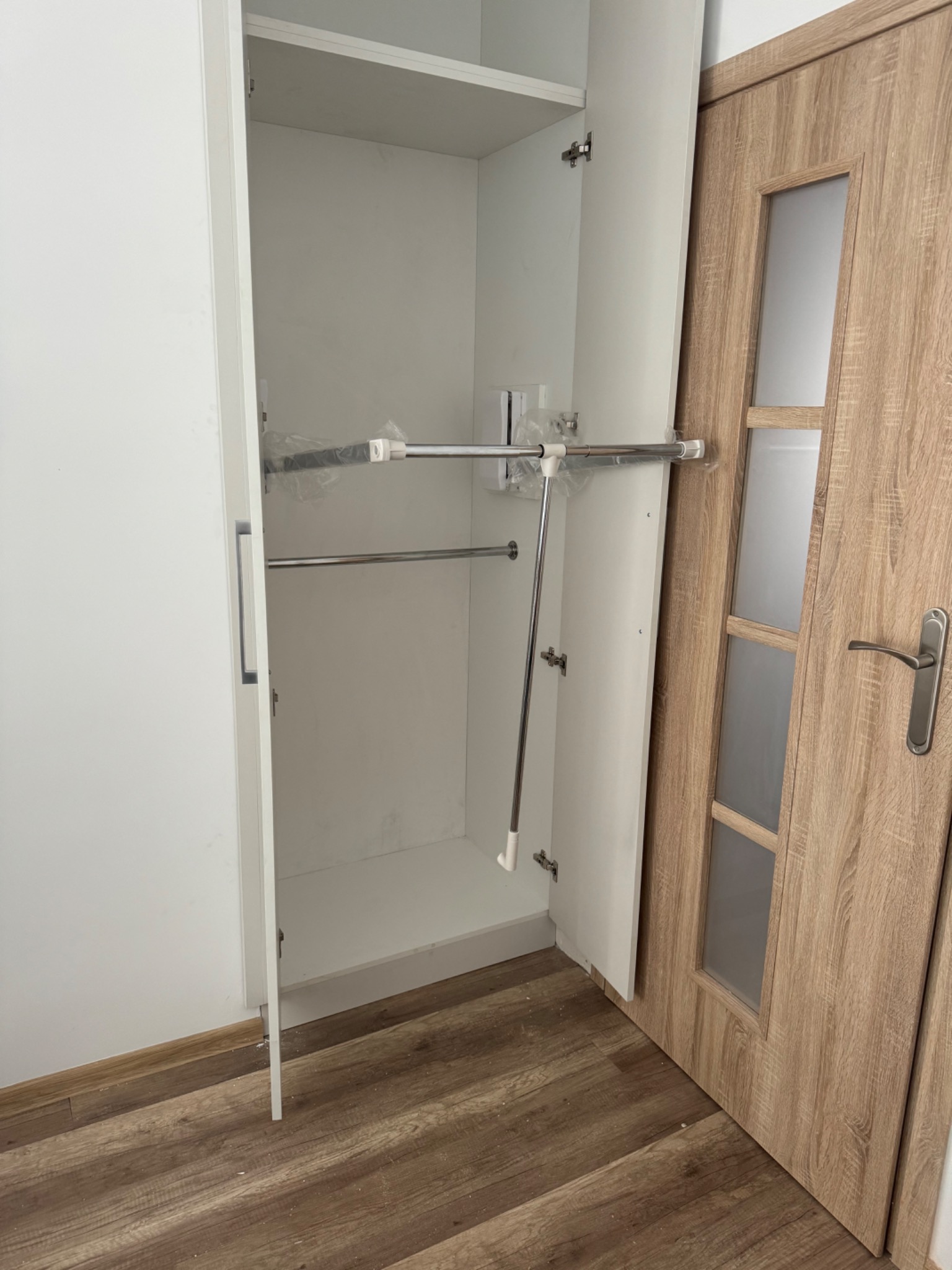 Wnętrze białej szafy wnękowej z zamontowanymi drążkami na ubrania, częściowo jeszcze zabezpieczonymi folią, widoczne na tle drewnopodobnej podłogi i drzwi.