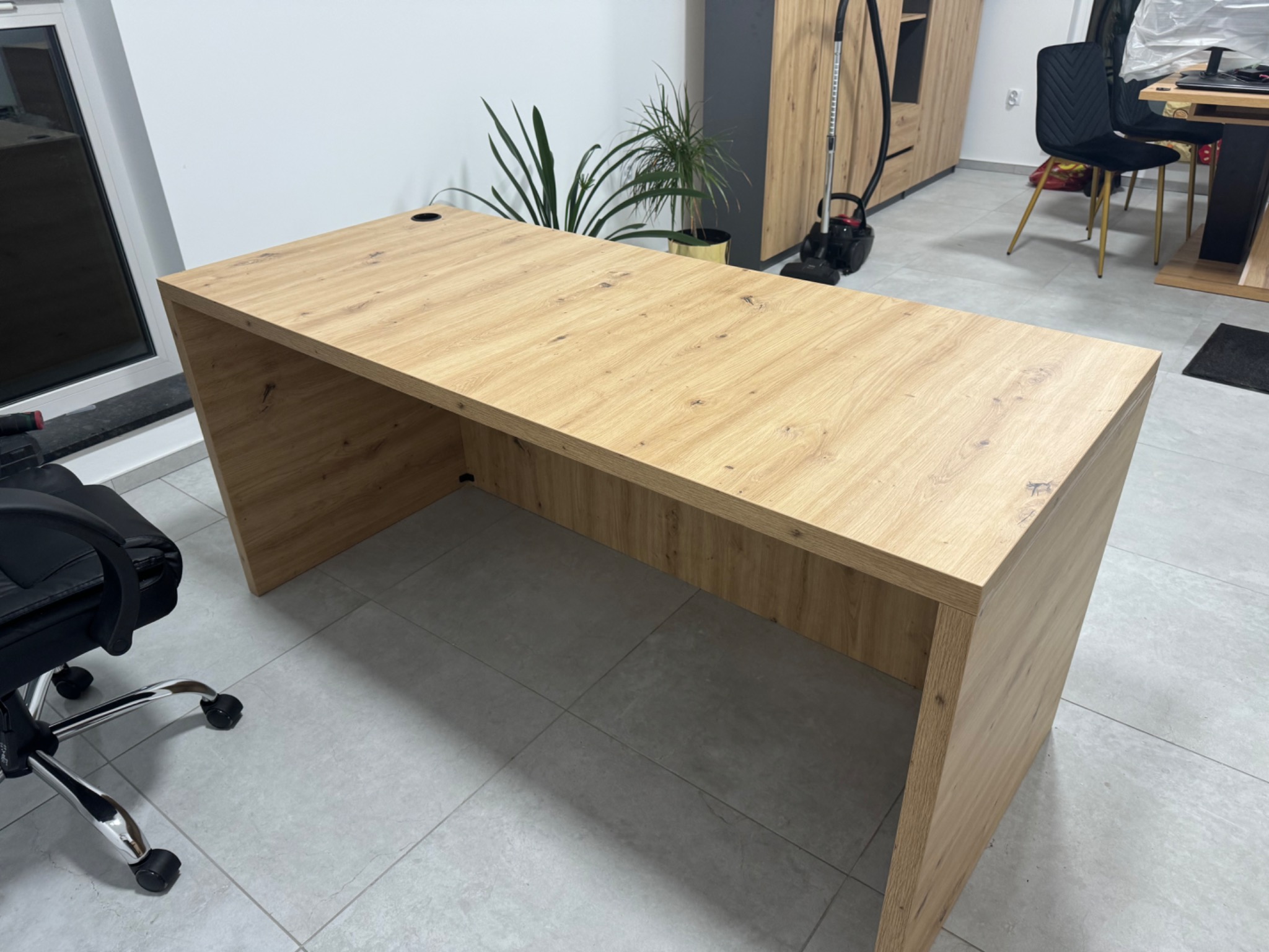 Biurko z płyty drewnopodobnej z otworem na kable, ustawione w pokoju z szarą podłogą i roślinami w tle.