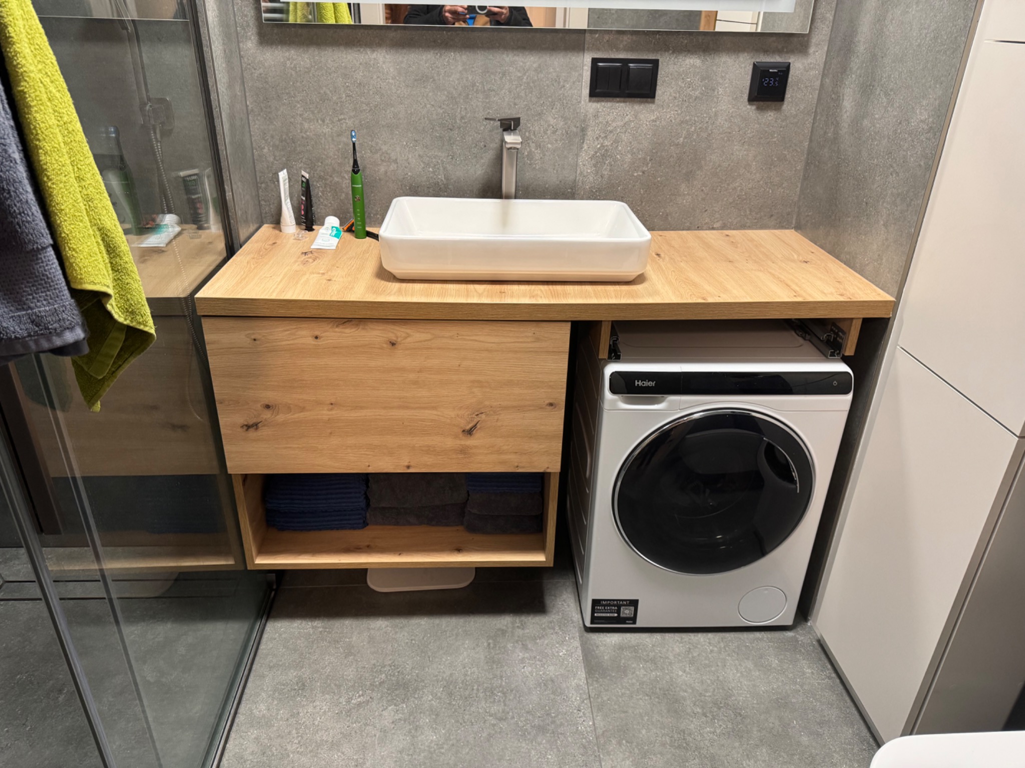Nowoczesna łazienka z drewnianym blatem pod umywalką, pod którym znajduje się półka z ręcznikami i pralka automatyczna.