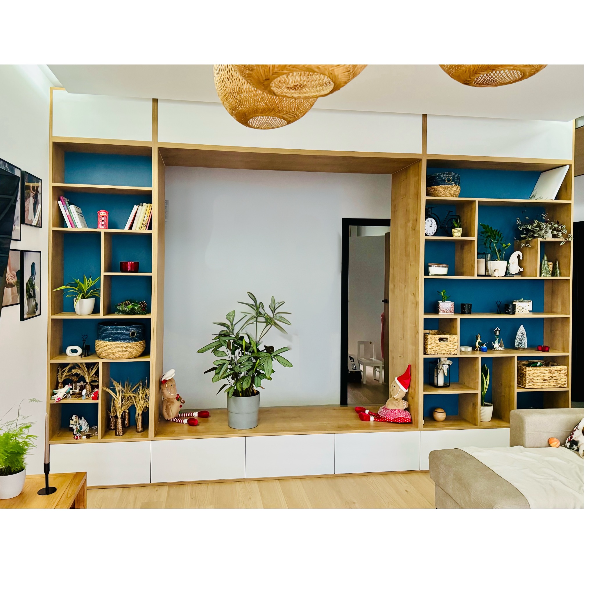 Duża, drewniana zabudowa meblowa z otwartymi półkami na tle błękitnej ściany, z dekoracjami i roślinami, uzupełniona białymi szafkami na dole, z widocznym fragmentem sofy i lampami z wikliny.