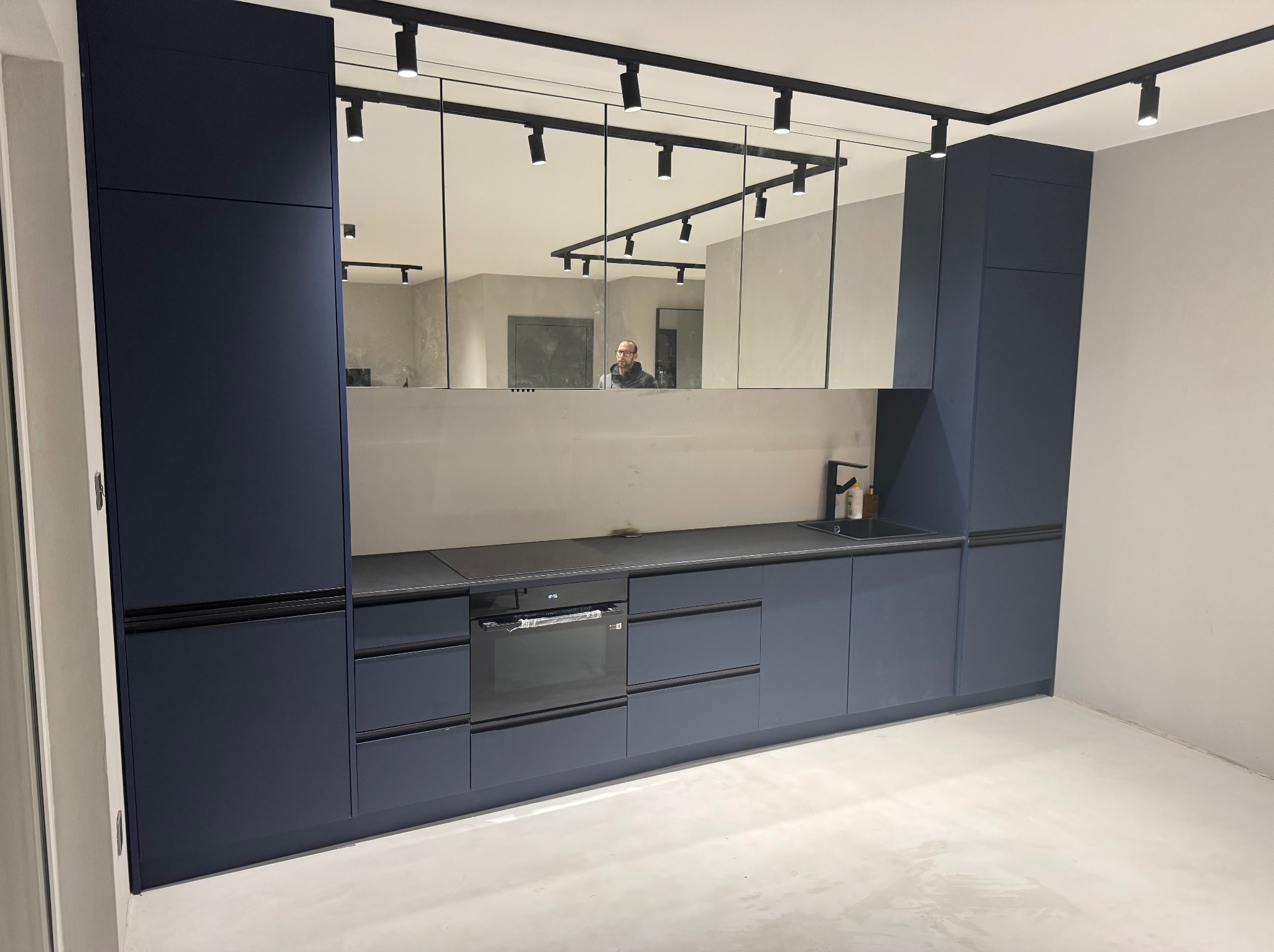 Nowoczesna kuchnia w odcieniach granatu z czarnym blatem i oświetleniem szynowym, odbicie osoby w lustrzanym panelu nad blatem.