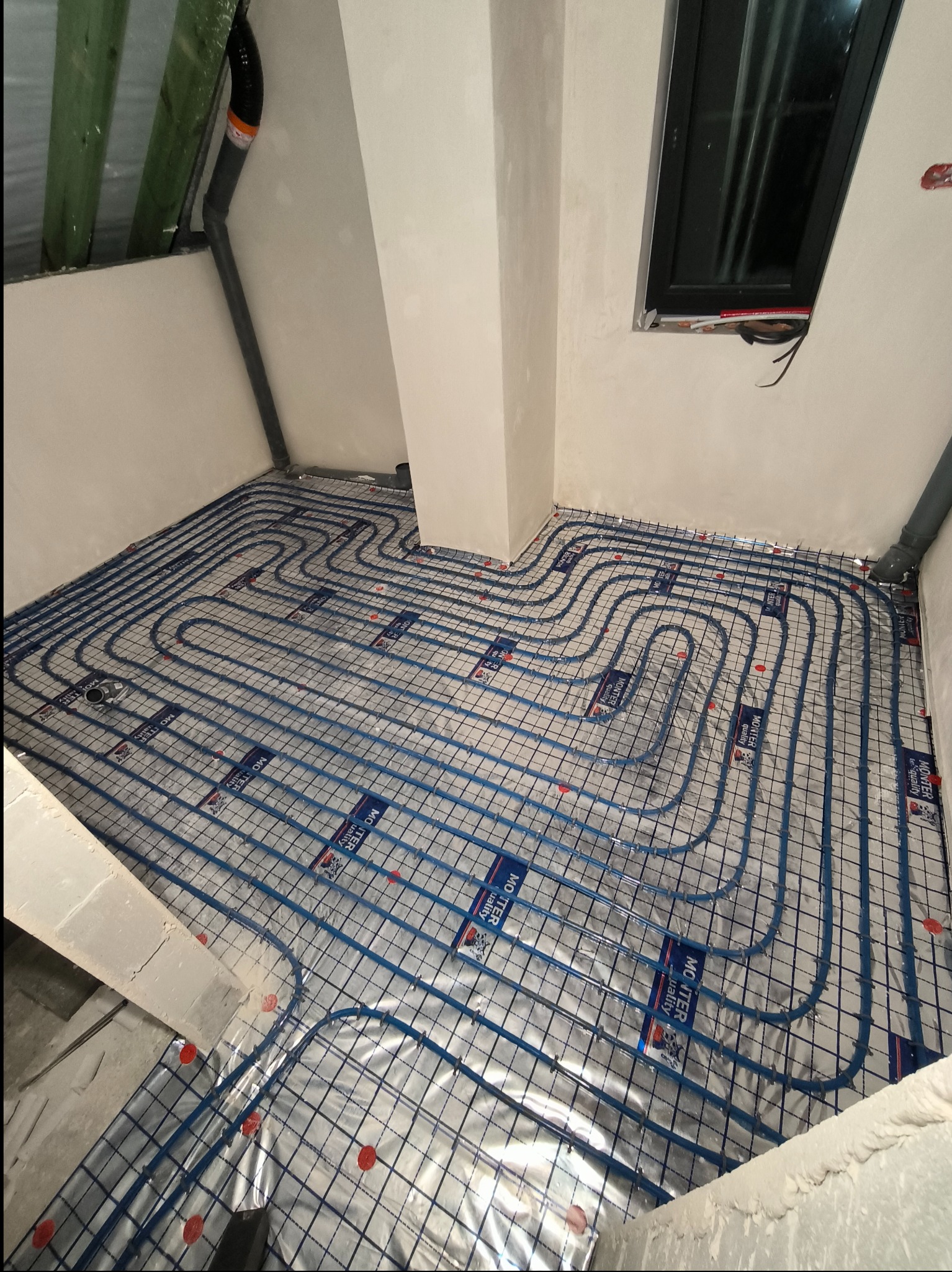 Układanie niebieskich rur ogrzewania podłogowego na folii izolacyjnej w pomieszczeniu na poddaszu, widoczne elementy konstrukcyjne dachu i pionowa rura kanalizacyjna.