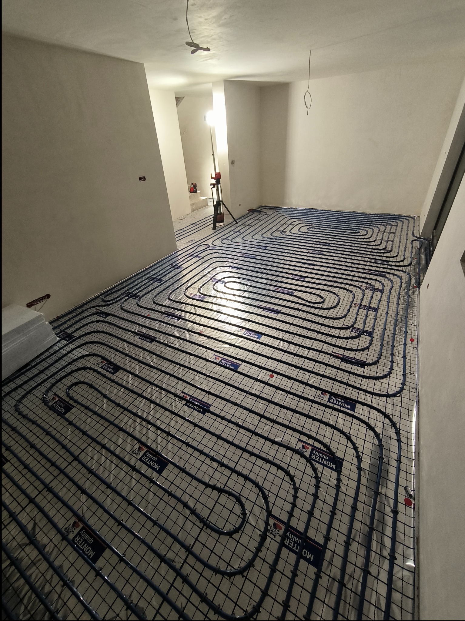 Rozłożona instalacja ogrzewania podłogowego w surowym wnętrzu, widoczne rury grzewcze ułożone w charakterystyczny wzór na folii izolacyjnej, w tle lampa budowlana oświetlająca dalszą część...