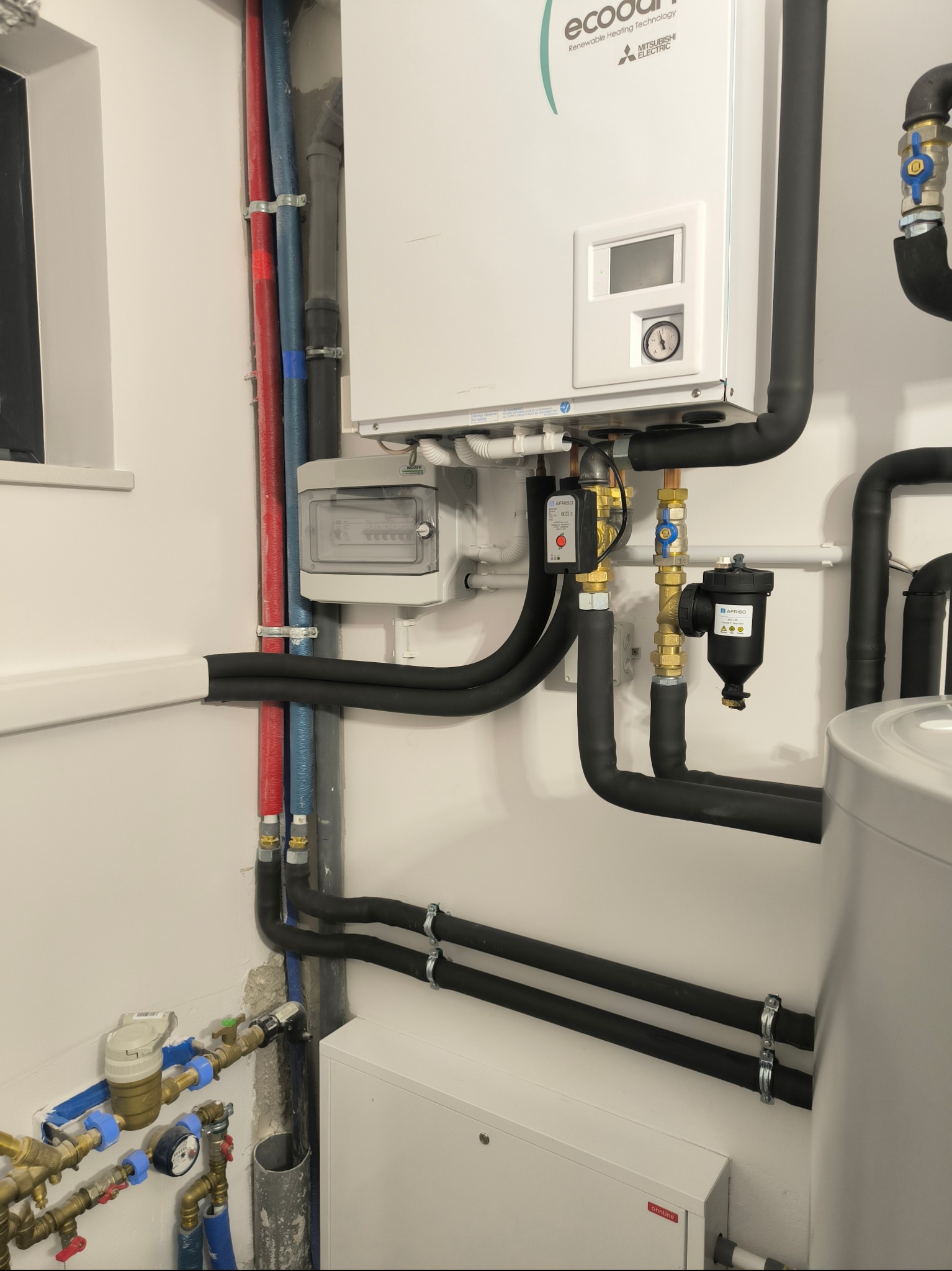 Nowoczesny piec gazowy z podłączeniami hydraulicznymi i elektrycznymi w schludnym pomieszczeniu technicznym, widoczne rury w izolacji, zawory, filtr oraz licznik wody.