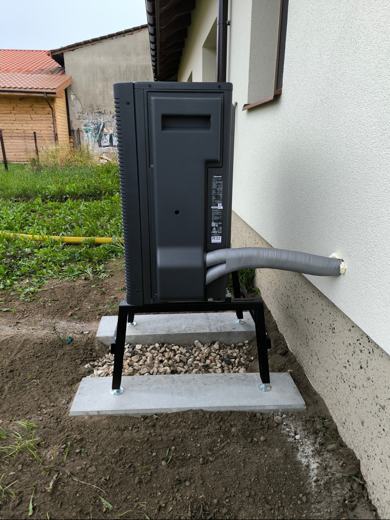 Zamontowana na zewnątrz budynku pompa ciepła Panasonic, na betonowej podstawie, z widocznymi przewodami w szarej izolacji wchodzącymi do ściany.