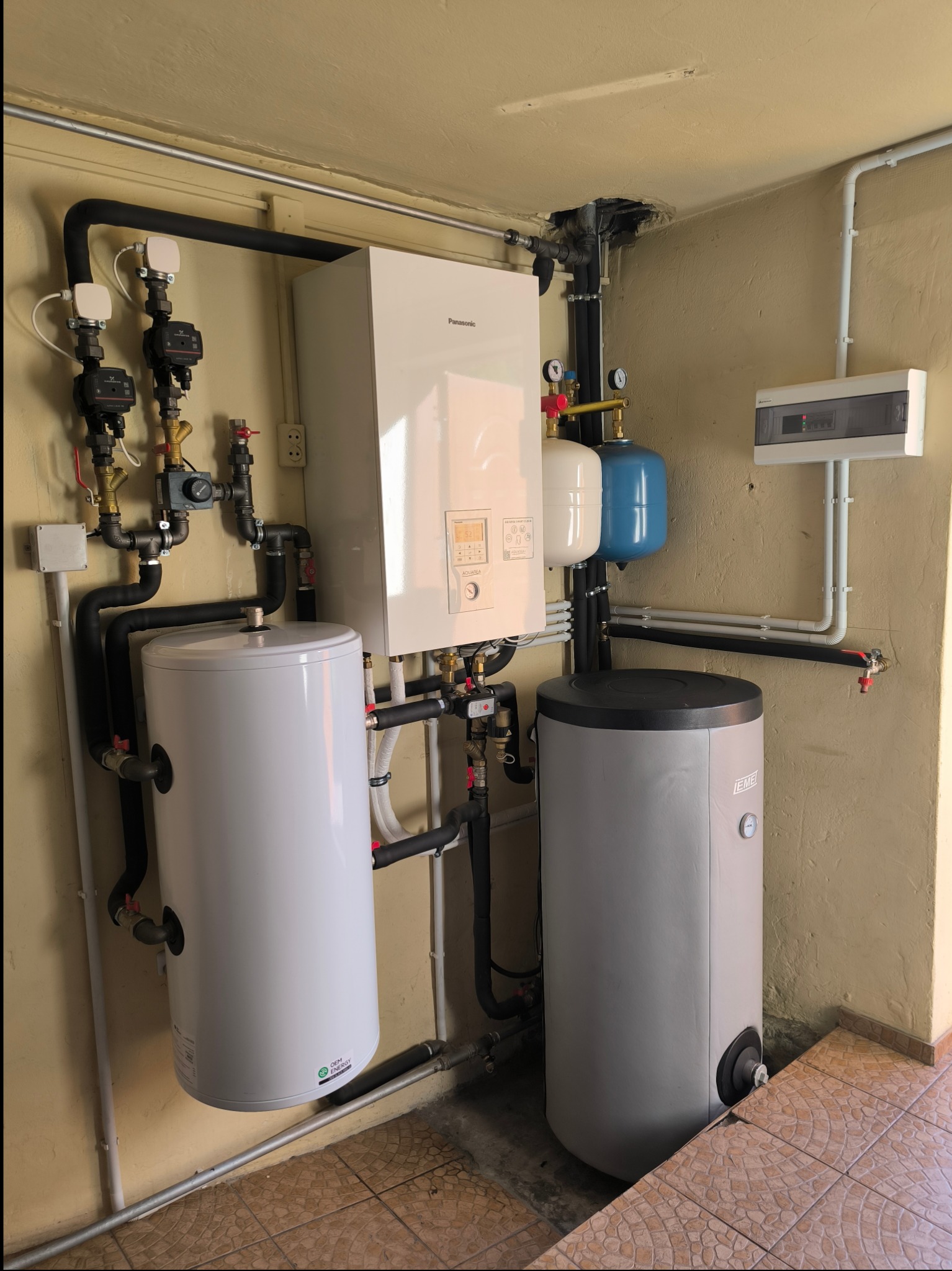 Biały piec gazowy Panasonic z dwoma zasobnikami wody (białym i szarym) oraz instalacją hydrauliczną w pomieszczeniu z beżowymi ścianami i brązową podłogą z płytek.