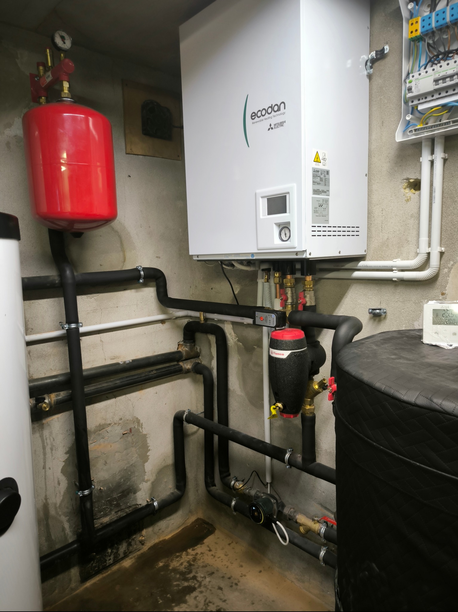 Zainstalowany piec CO marki Ecodan z podłączonymi rurami, zbiornikiem oraz rozdzielnicą elektryczną na surowej ścianie w pomieszczeniu technicznym.