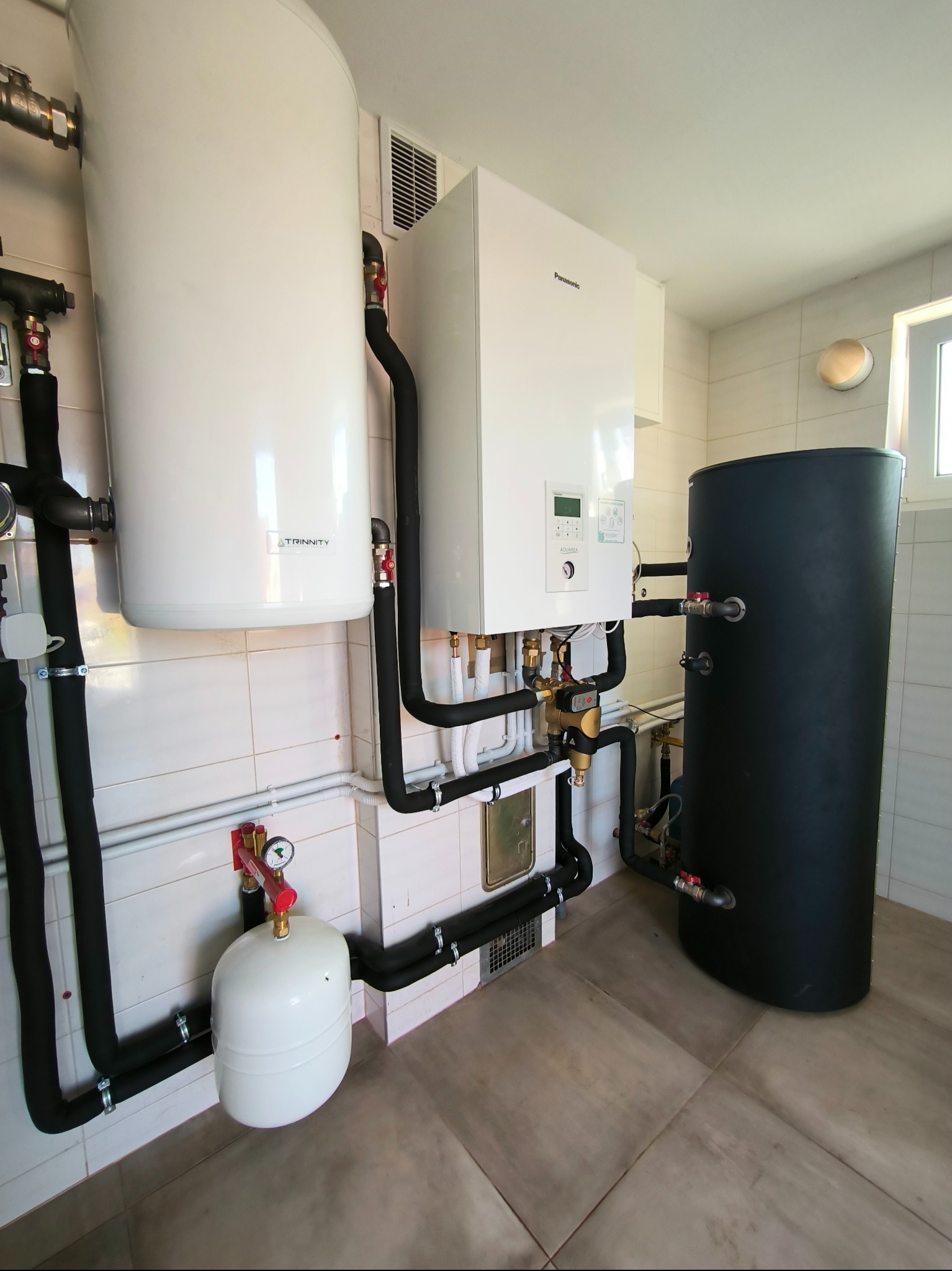 Kotłownia z piecem gazowym Panasonic, zasobnikiem ciepłej wody Trinity i czarnym zbiornikiem akumulacyjnym, połączone rurami w izolacji, na tle białych płytek ściennych i beżowej podłogi.