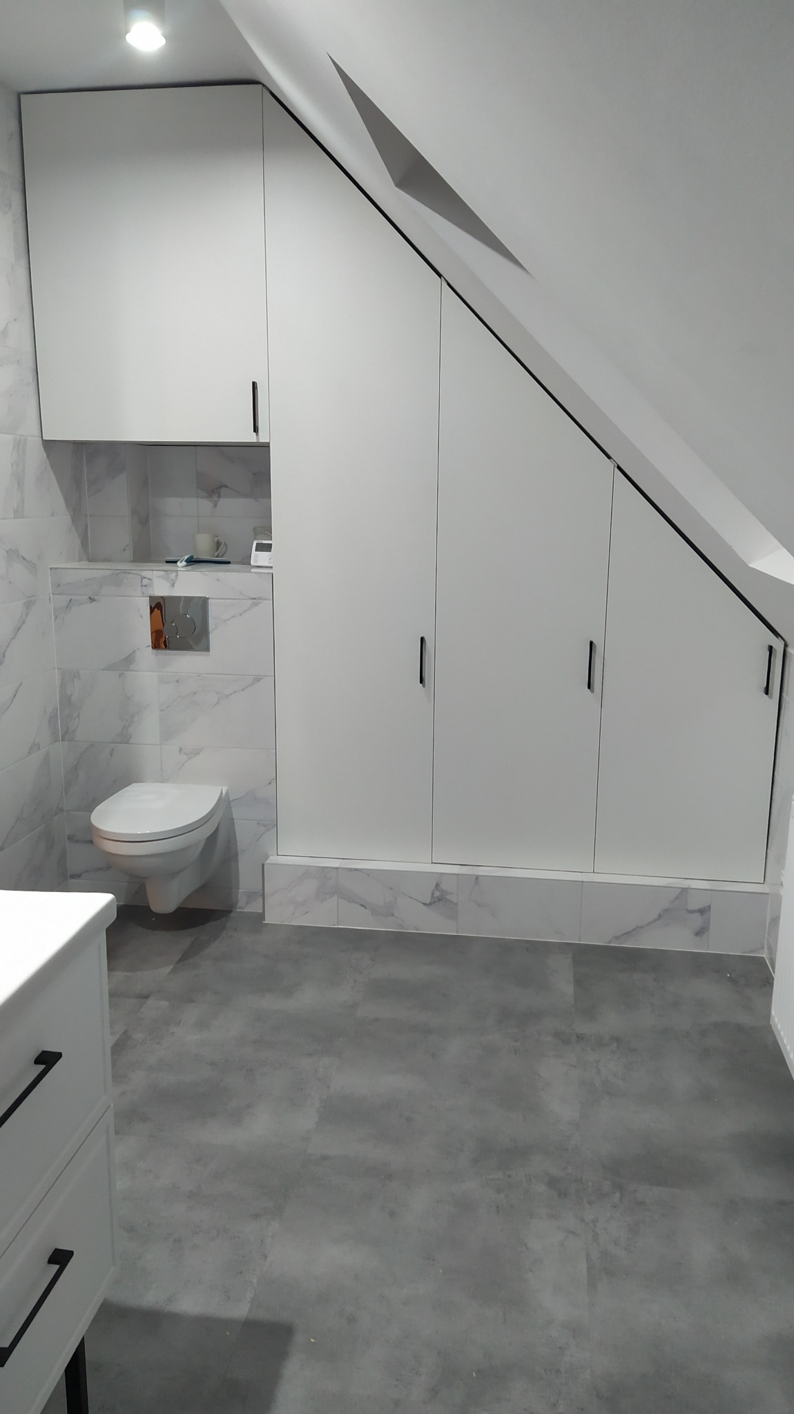 Biała zabudowa pod skosem dachu z czarnymi uchwytami w łazience z toaletą i marmurowymi płytkami na ścianach.