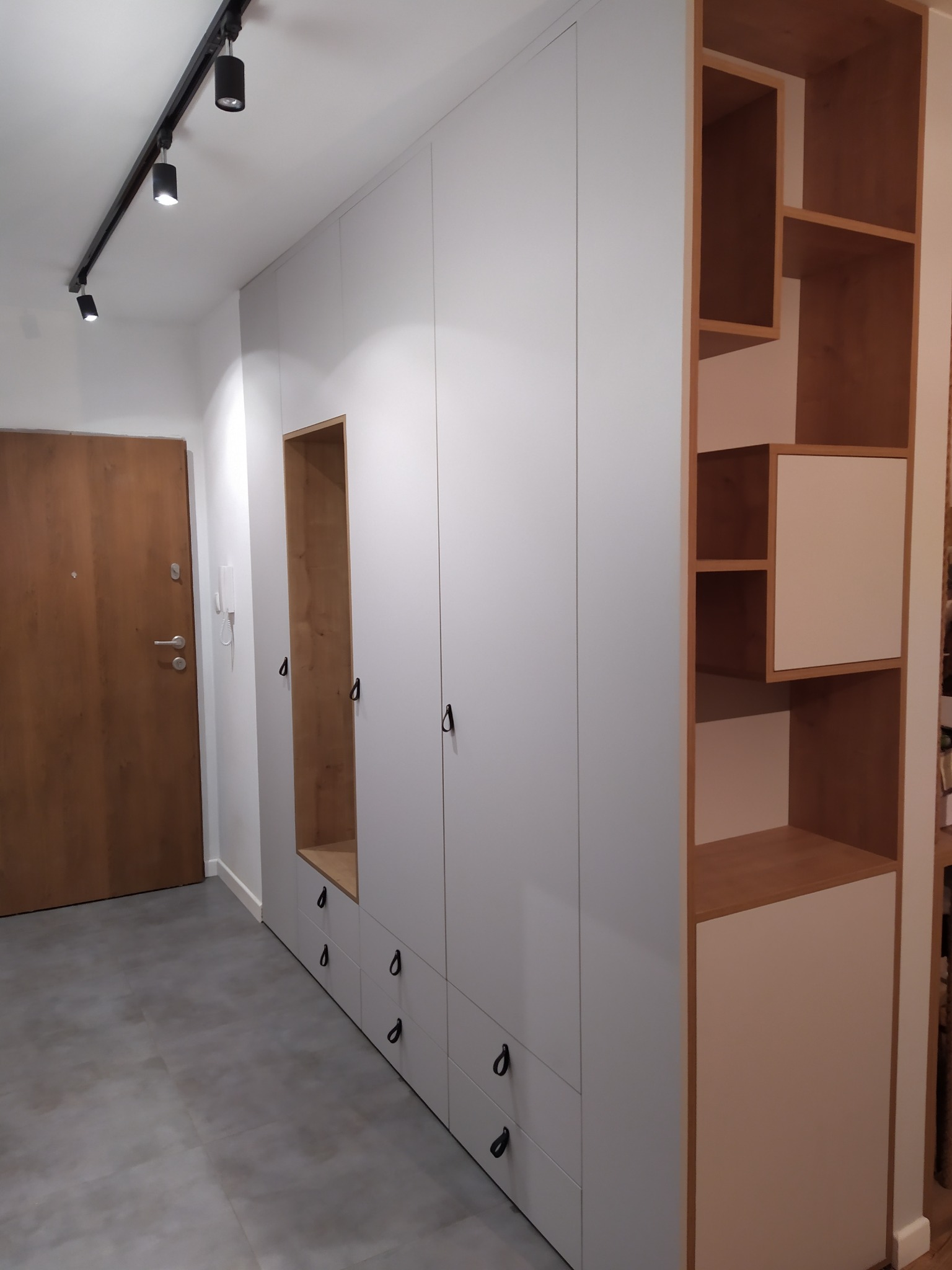 Biała szafa wnękowa z drewnianymi akcentami i otwartymi półkami w przedpokoju. Minimalistyczny design, czarne uchwyty, oświetlenie szynowe.