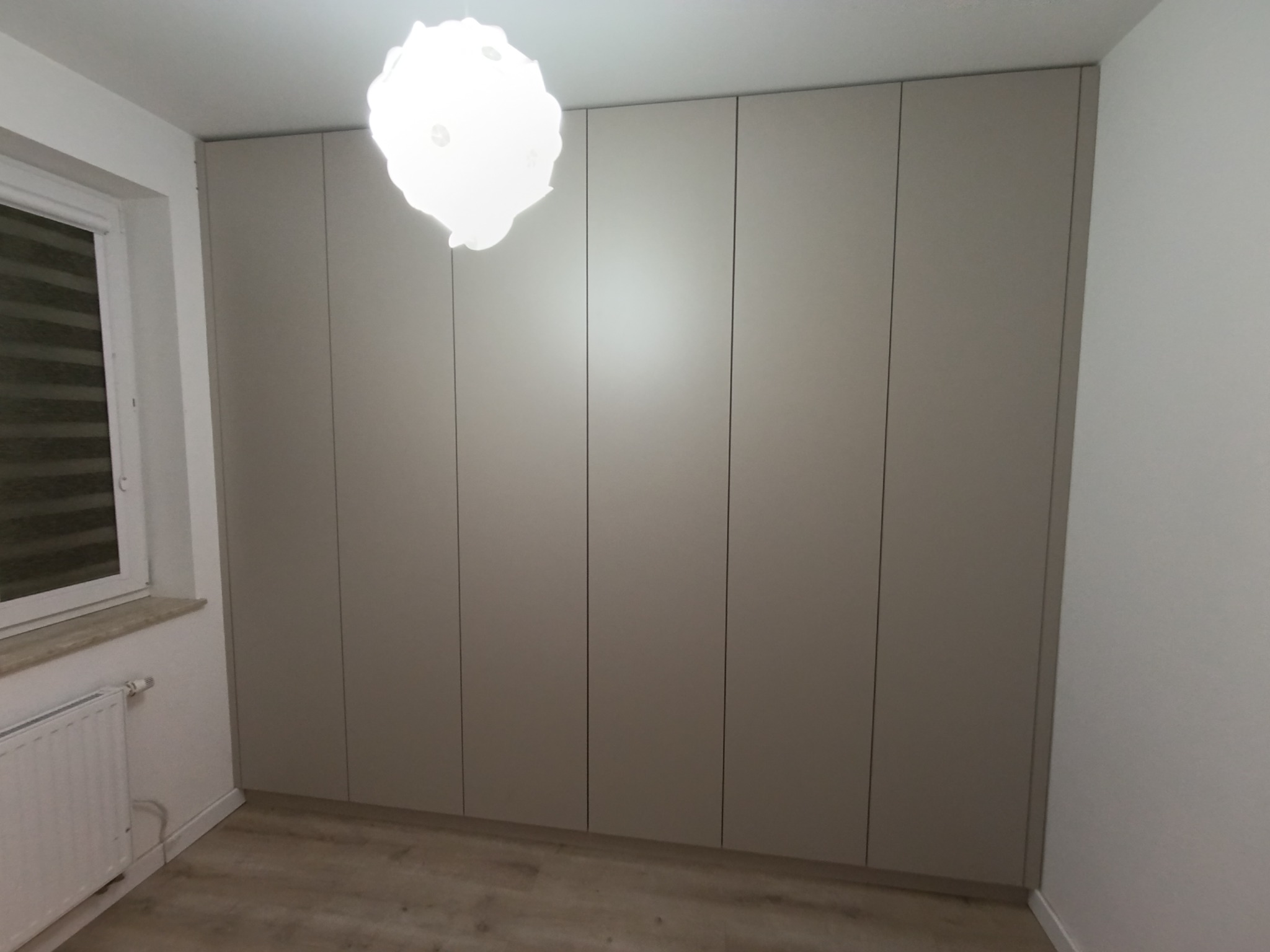 Beżowa szafa wnękowa z sześcioma drzwiami, dopasowana do ściany, z widocznym oknem i grzejnikiem. Minimalistyczny design, jasne wnętrze.