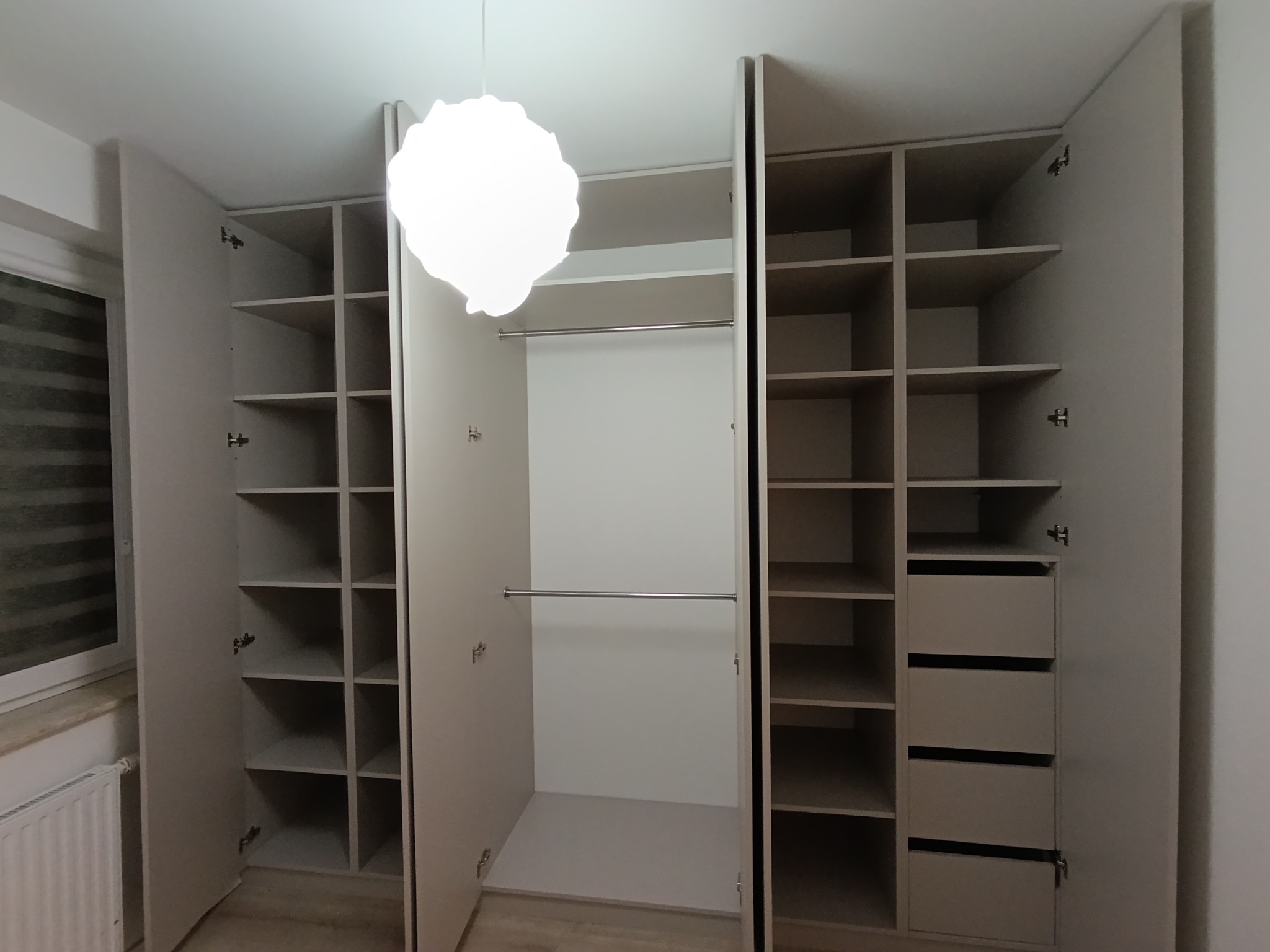 Jasna szafa wnękowa z otwartymi drzwiami, widoczne półki, drążki i szuflady. Minimalistyczny design, oświetlenie sufitowe. Widok z przodu, wnętrze szafy.