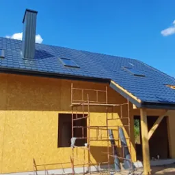 Nowy dom w trakcie budowy, z ciemnoszarą dachówką, oknami dachowymi i kominem, ściany zewnętrzne z płyt OSB, widoczne rusztowanie.