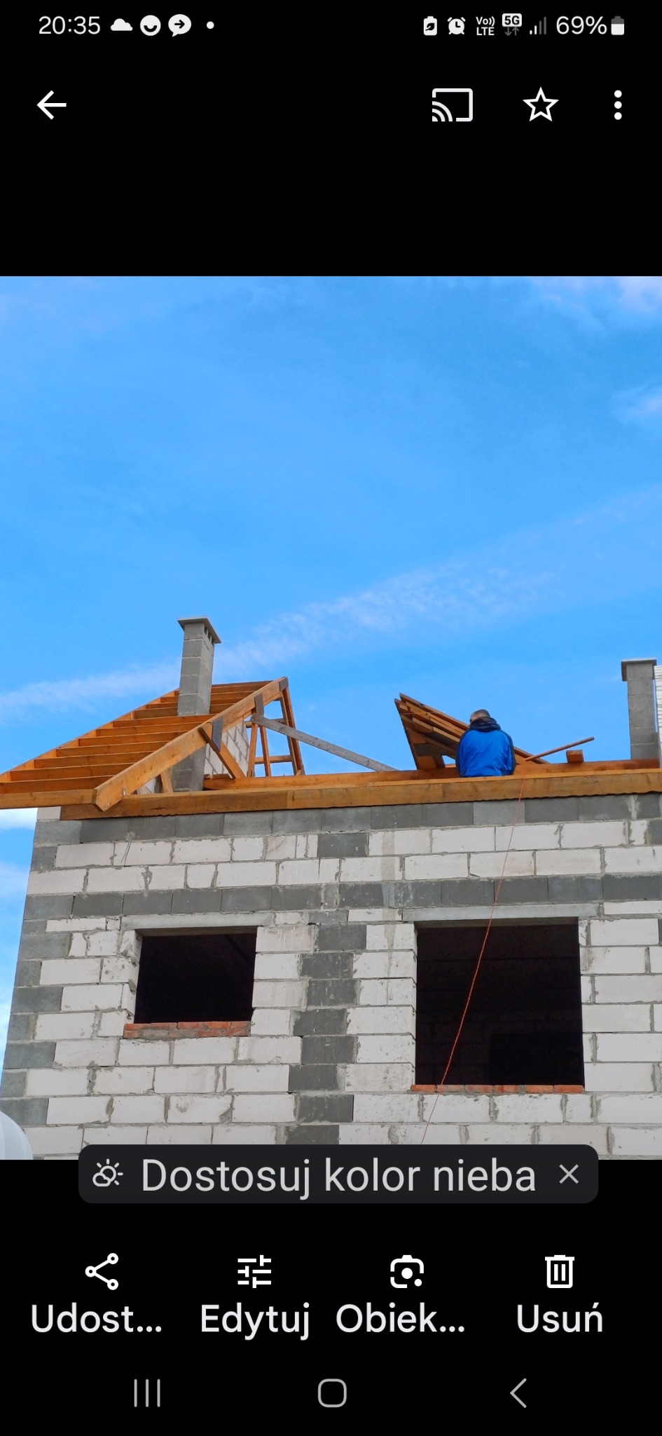 Montaż drewnianej konstrukcji dachu na budynku z szarego bloczka, widoczny komin i pracownik w niebieskiej kurtce oraz błękitne niebo w tle.