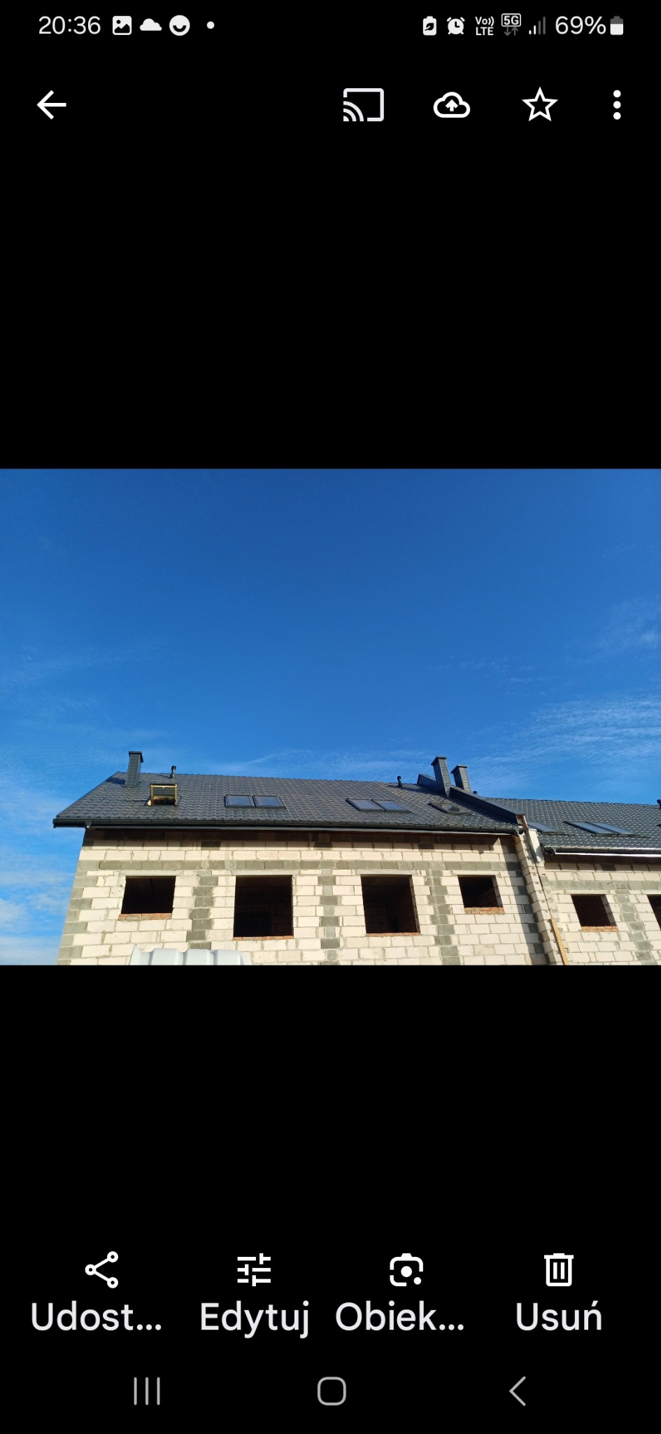 Surowy budynek z szarego bloczka z nowym, ciemnym dachem i oknami dachowymi na tle błękitnego nieba.