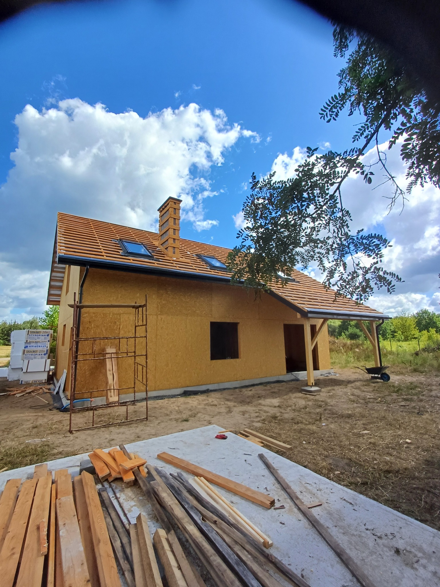 Dom w trakcie budowy z widocznym dachem pokrytym dachówką, kominem i oknami dachowymi, elewacja z płyt OSB, fragment rusztowania, deski na pierwszym planie, błękitne niebo z chmurami w tle.
