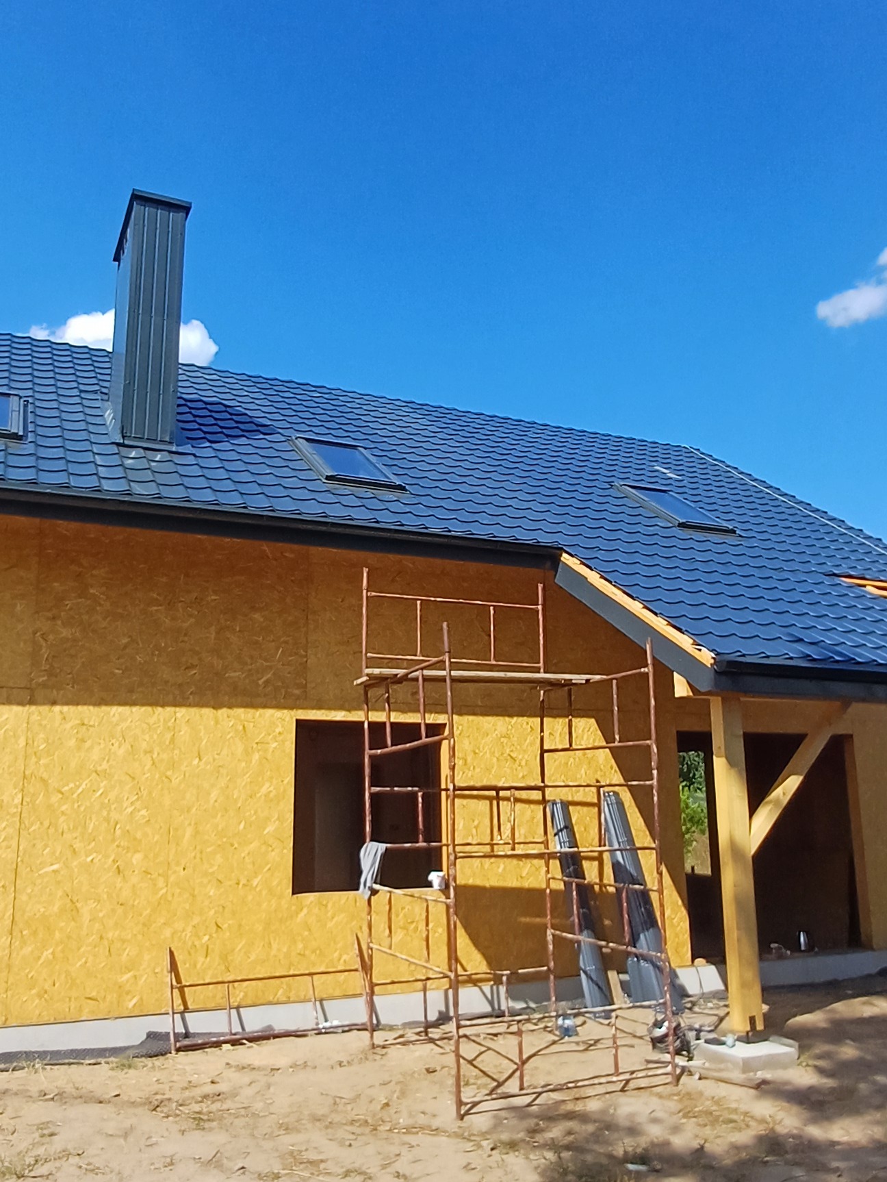 Nowy dom w trakcie budowy, z ciemnoszarą dachówką, oknami dachowymi i kominem, ściany zewnętrzne z płyt OSB, widoczne rusztowanie.