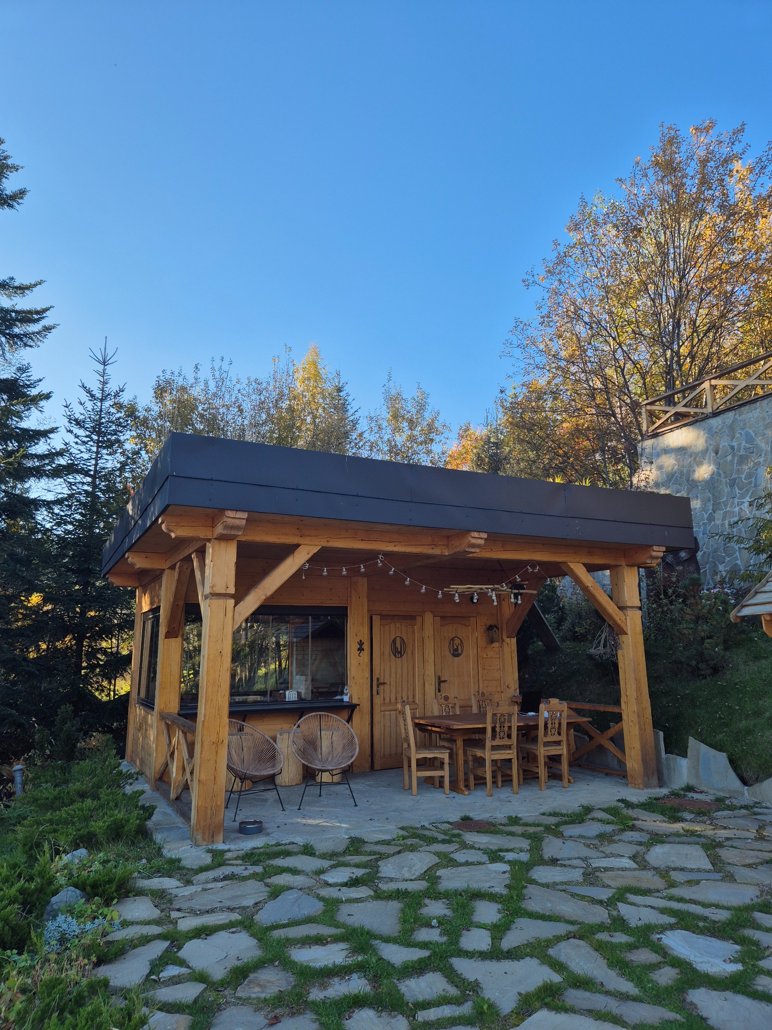 Drewniana altana z płaskim dachem i oświetleniem sznurkowym, otoczona drzewami i kamienną ścieżką z rosnącą trawą, widok na błękitne niebo.