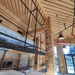 FOREST SPIRIT - Wnętrze w stylu rustykalnym z drewnianym stropem, stalową konstrukcją podtrzymującą, kamiennym filarem, widokiem na góry na ścianie, oświetleniem w stylu retro i częściowo zamontowaną zabudową...