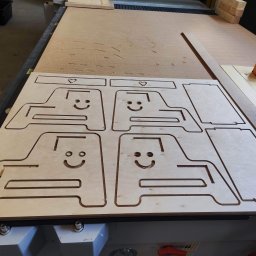 FOREST SPIRIT - Płyta ze sklejki z wyciętymi za pomocą plotera CNC czterema identycznymi elementami w kształcie stylizowanych samochodów z uśmiechniętymi buziami i serduszkami, leżąca na stole roboczym.