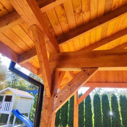 FOREST SPIRIT - Drewniana konstrukcja z widocznym dachem, belkami i słupami, z rynną odprowadzającą wodę, w tle zielony trawnik, wysokie tuje i domek dla dzieci ze zjeżdżalnią.