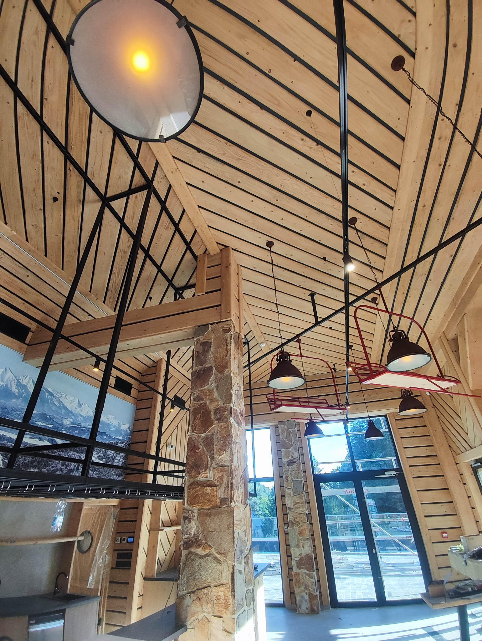 Wnętrze budynku z drewnianą konstrukcją dachu, kamiennymi filarami i oświetleniem w stylu loft, widok na góry przez okno.