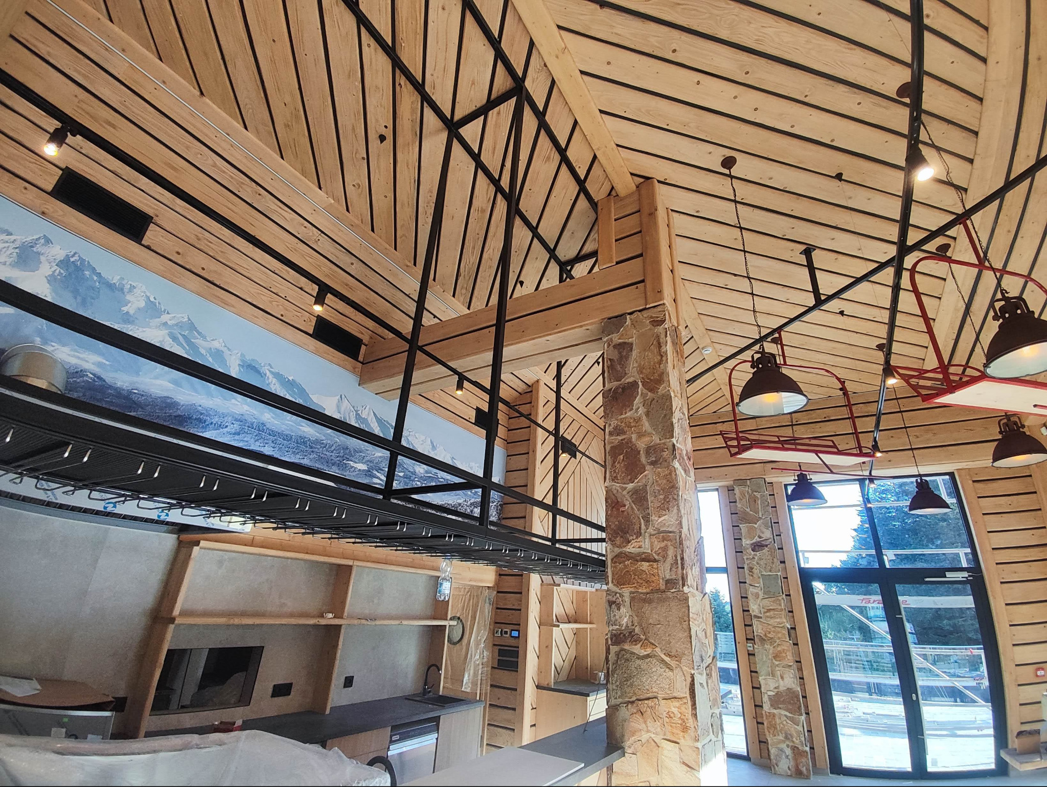 Wnętrze w stylu rustykalnym z drewnianym stropem, stalową konstrukcją podtrzymującą, kamiennym filarem, widokiem na góry na ścianie, oświetleniem w stylu retro i częściowo zamontowaną zabudową...