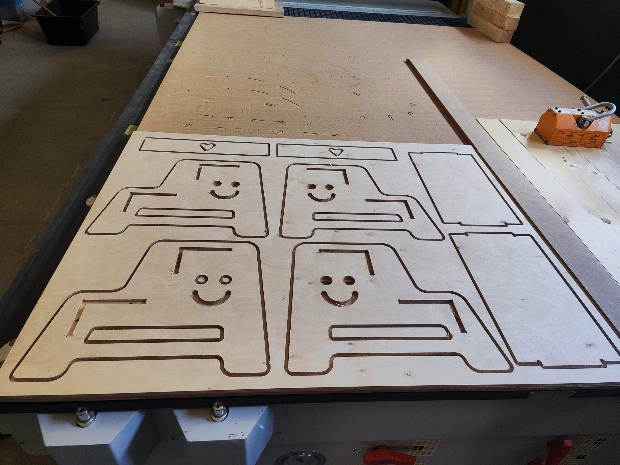 Płyta ze sklejki z wyciętymi za pomocą plotera CNC czterema identycznymi elementami w kształcie stylizowanych samochodów z uśmiechniętymi buziami i serduszkami, leżąca na stole roboczym.