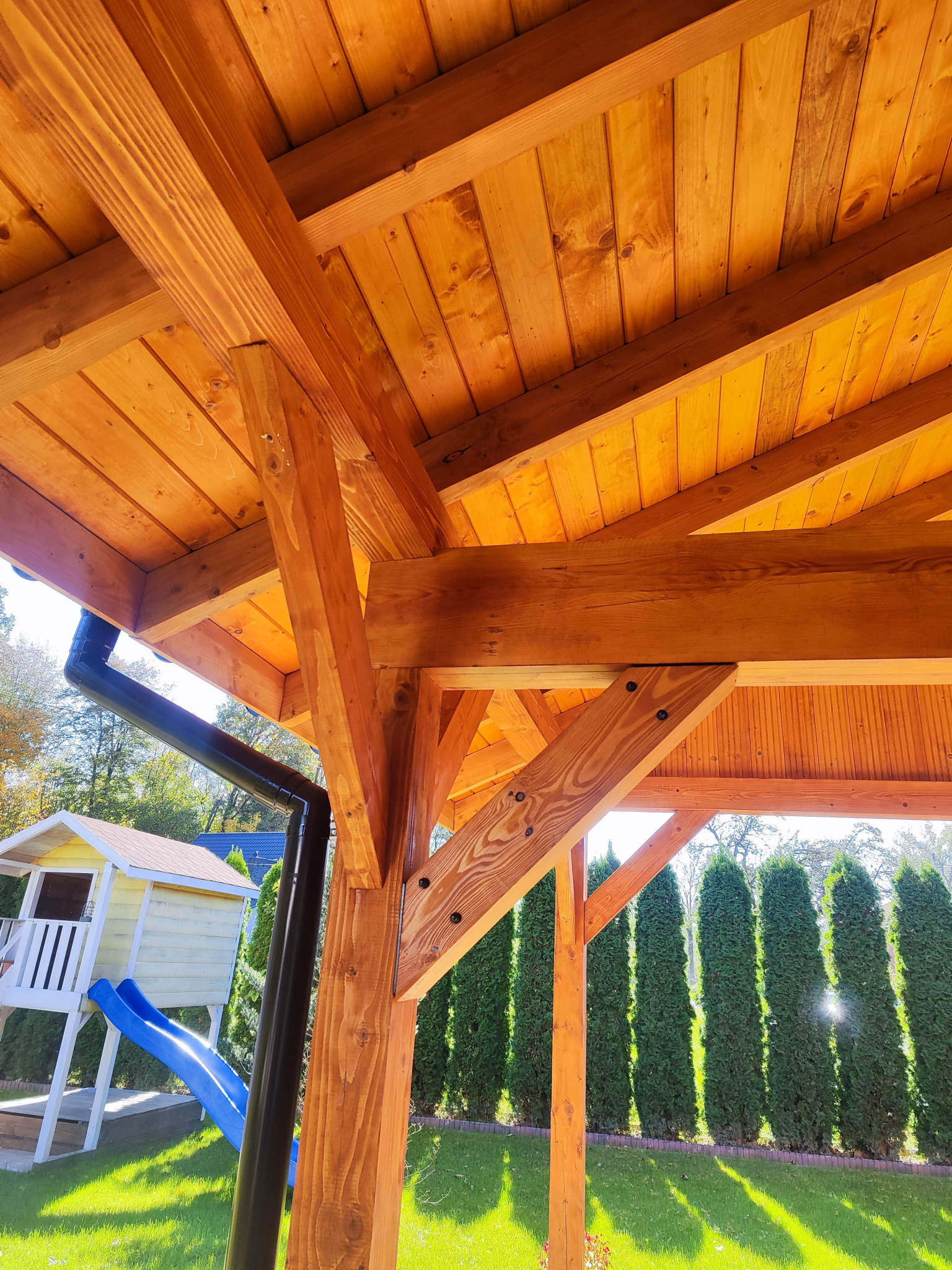 Drewniana konstrukcja z widocznym dachem, belkami i słupami, z rynną odprowadzającą wodę, w tle zielony trawnik, wysokie tuje i domek dla dzieci ze zjeżdżalnią.