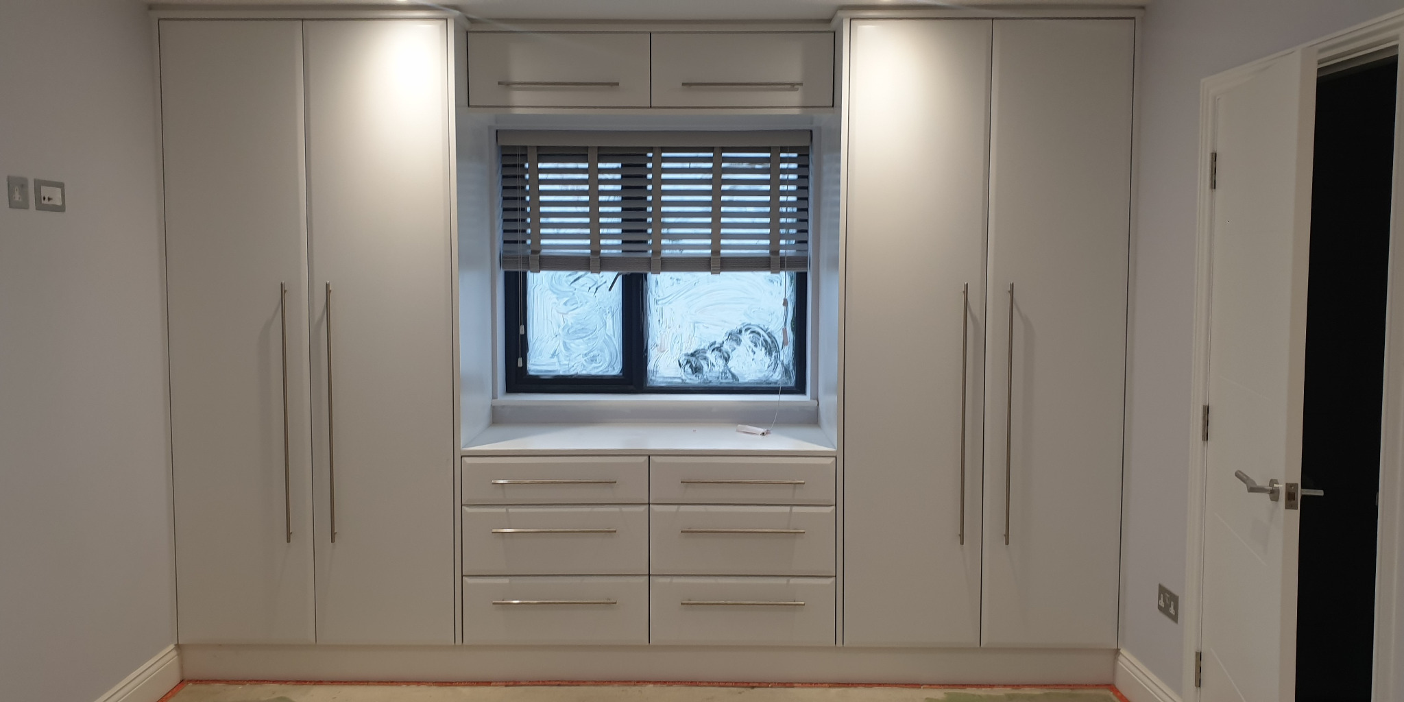 Biała zabudowa meblowa z szafami po bokach okna z żaluzjami, uzupełniona o szafki z szufladami poniżej parapetu.