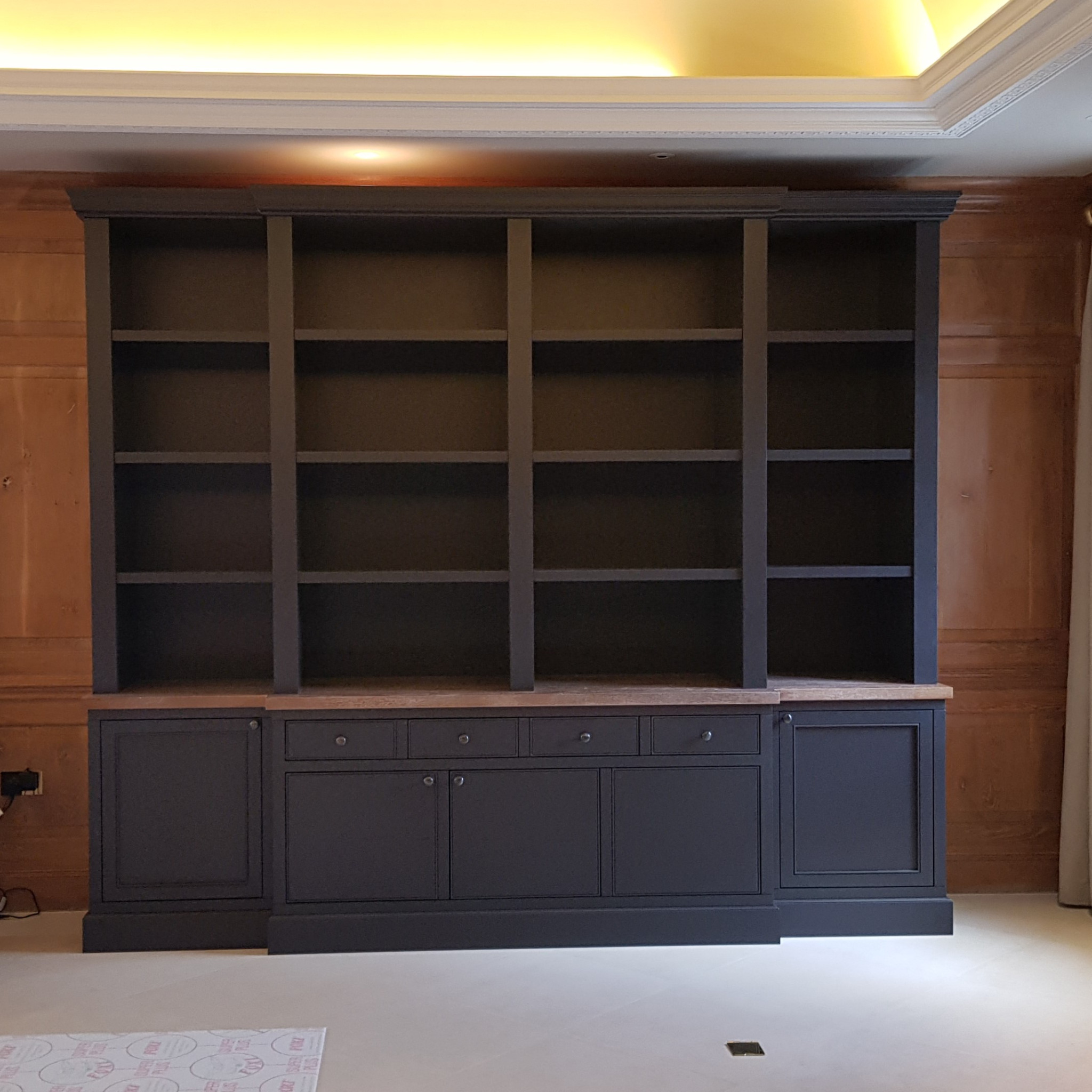 Czarna, zabudowana biblioteczka z otwartymi półkami i szafkami na dole, zamontowana przy ścianie z drewnianą boazerią.