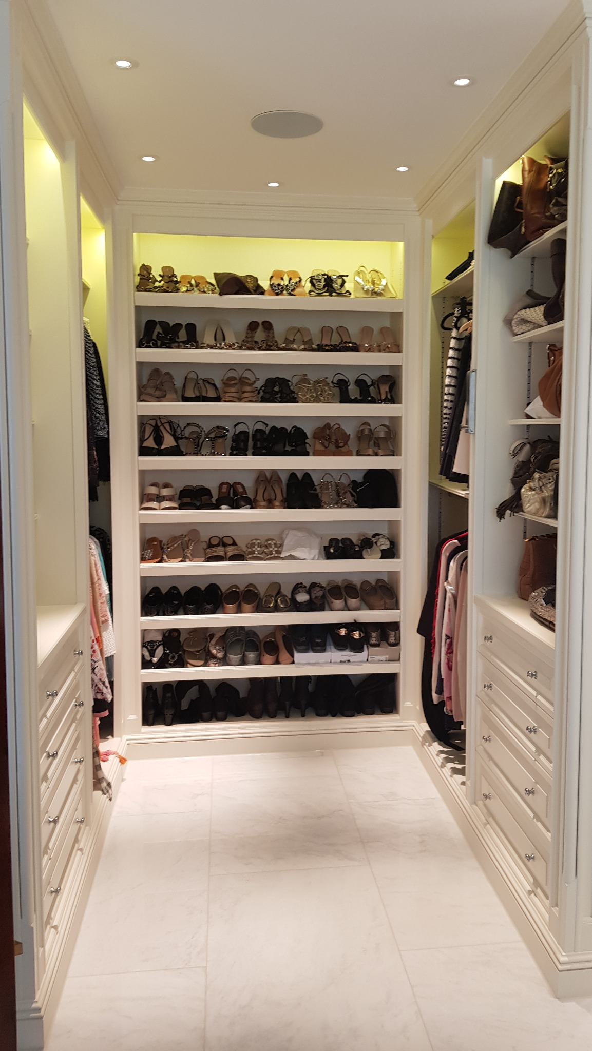 Wnętrze garderoby z białymi szafami na wymiar, półkami pełnymi butów i ubrań, oświetleniem punktowym.