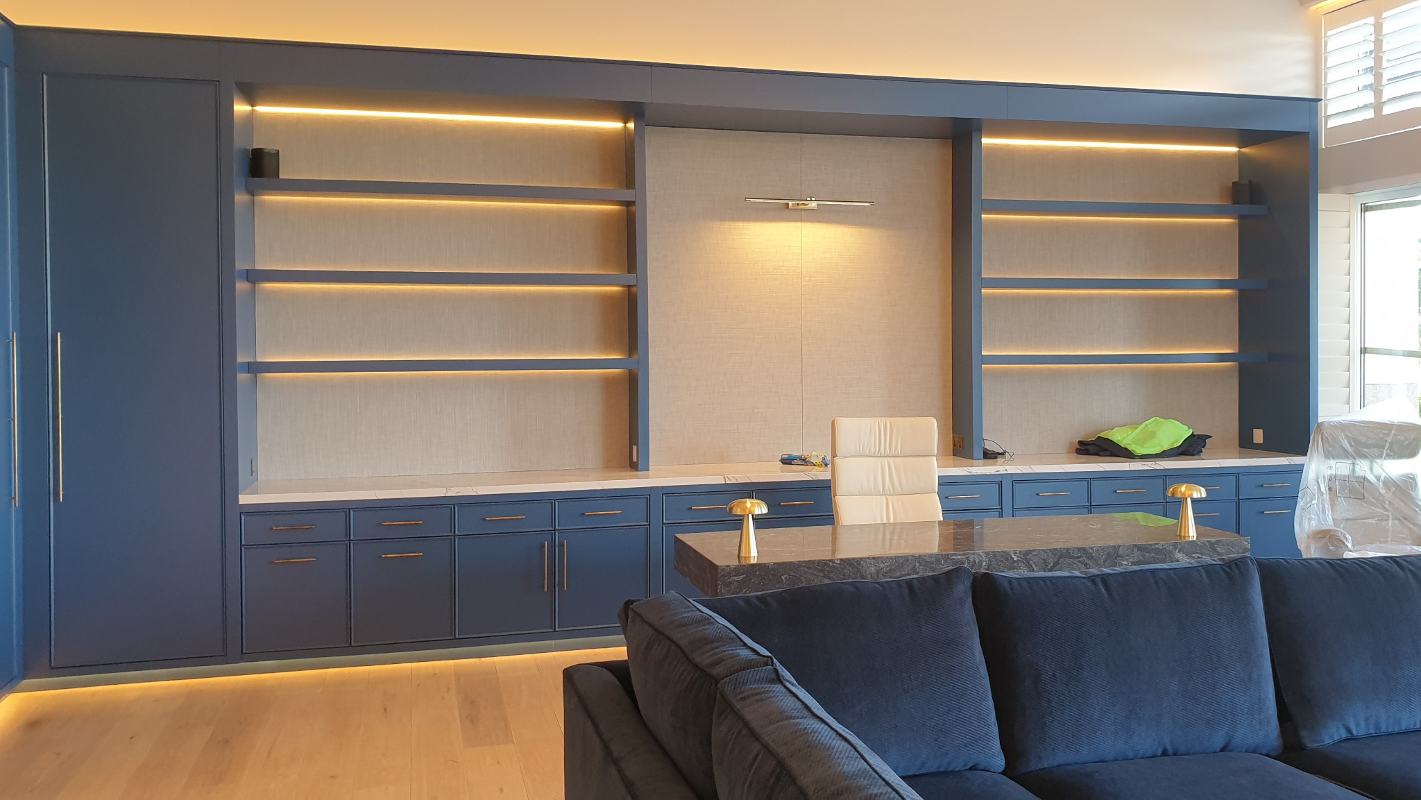 Niebieska zabudowa meblowa z podświetlanymi półkami, biurkiem z marmurowym blatem i krzesłem biurowym, widok zza sofy w salonie.