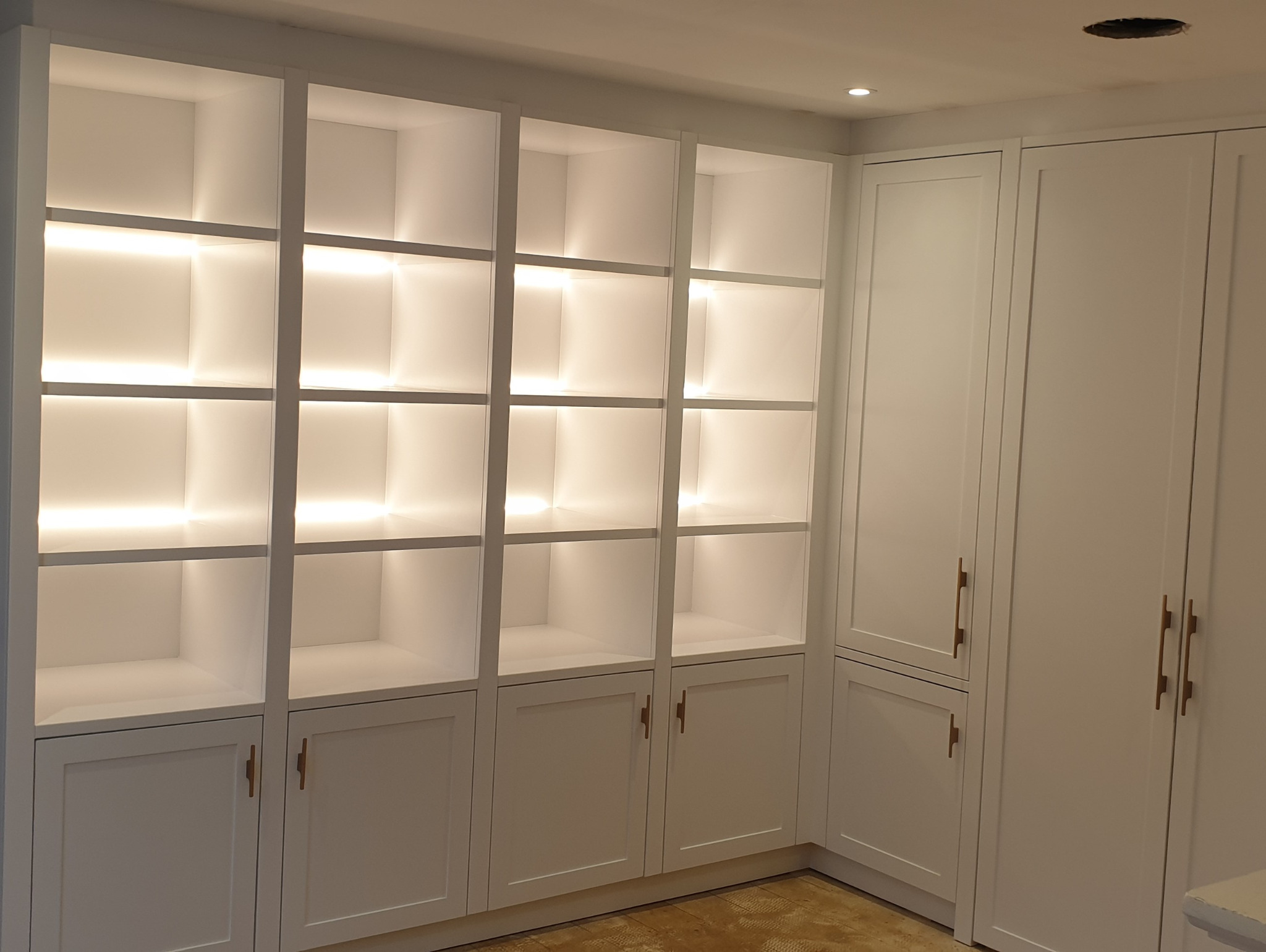 Biała zabudowa meblowa z podświetlanymi półkami i szafkami, minimalistyczne uchwyty, zabudowa do sufitu.