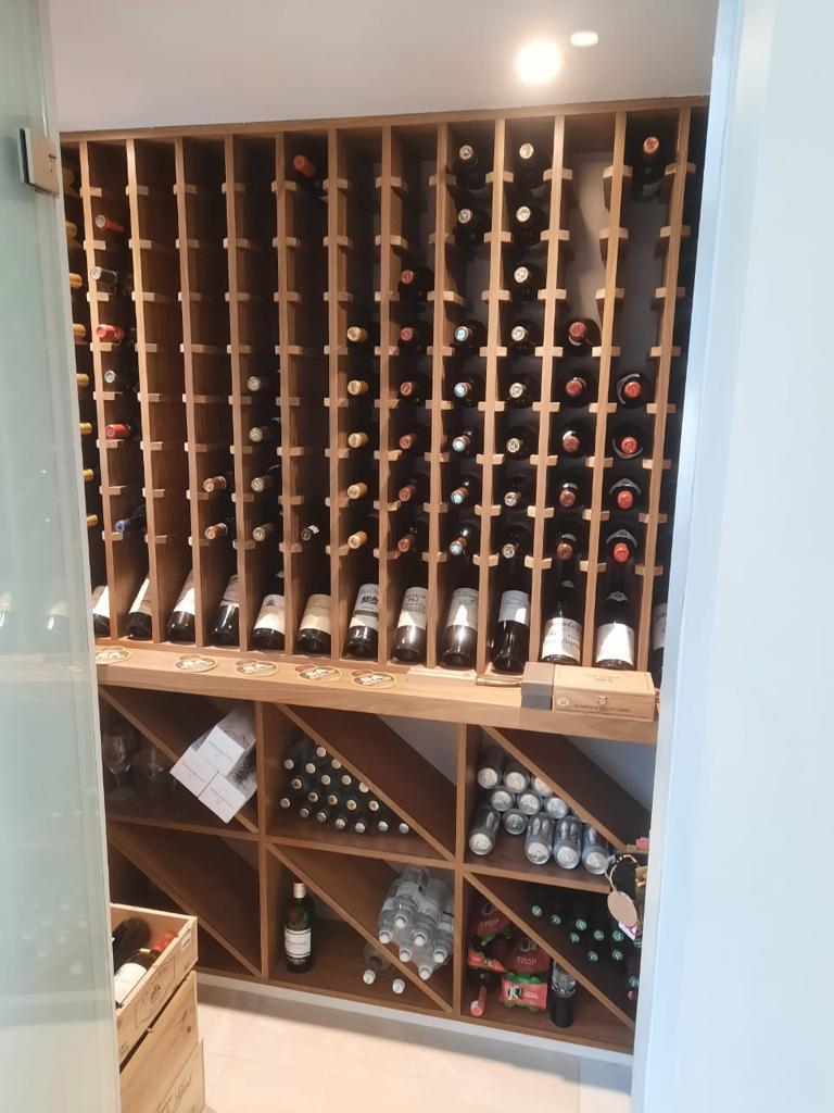 Drewniana zabudowa wnęki z półkami na wino i inne napoje, dolna część z ukośnymi przegródkami, oświetlenie punktowe u góry.