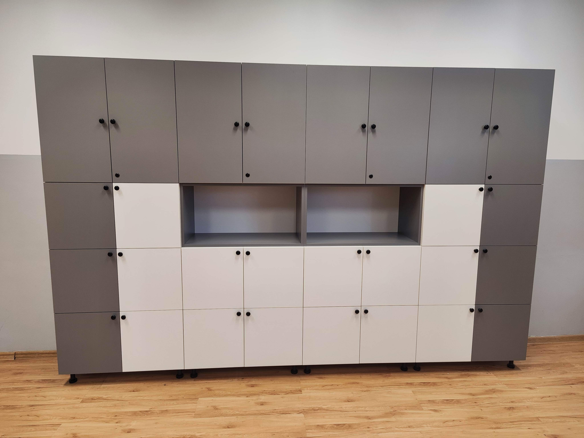 Duża, modułowa szafa z szarymi i białymi frontami, wyposażona w czarne, okrągłe uchwyty, umieszczona na kółkach, stojąca na drewnianej podłodze w jasnym pomieszczeniu.