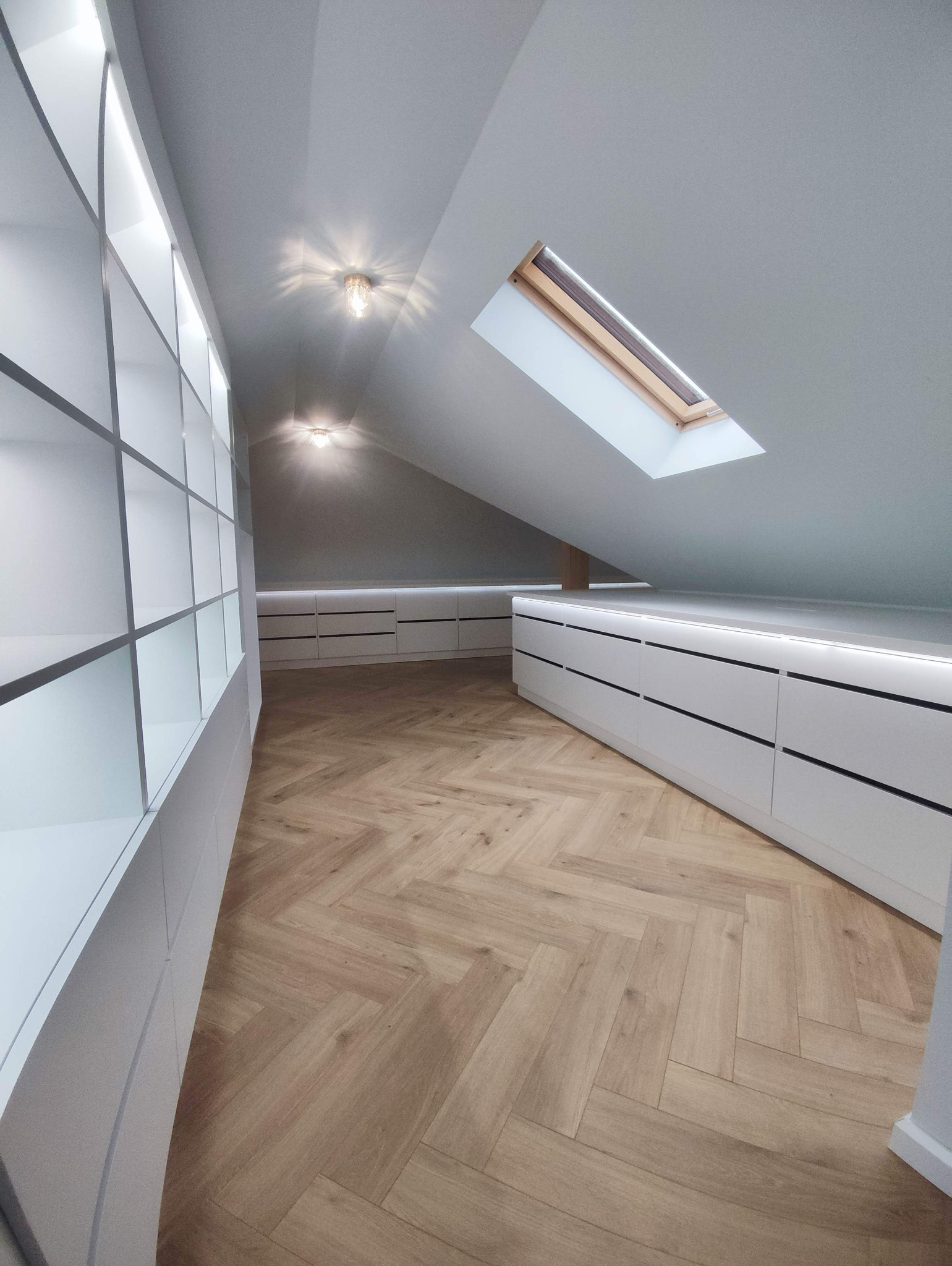 Garderoba na poddaszu ze skośnym sufitem, oświetleniem LED i parkietem ułożonym we wzór jodełki.
