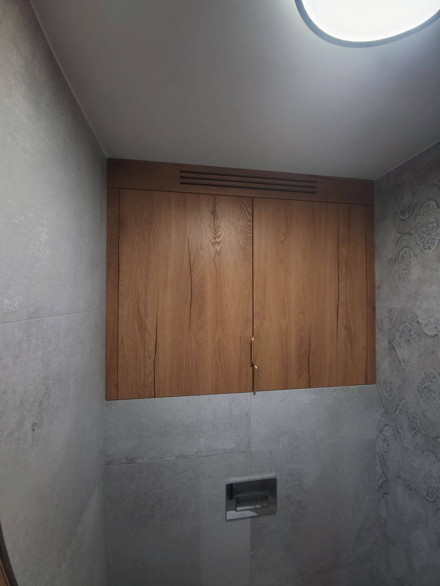 Drewniana szafka łazienkowa wisząca nad toaletą, z widocznymi uchwytami i wentylacją, na tle szarych płytek z wzorem.