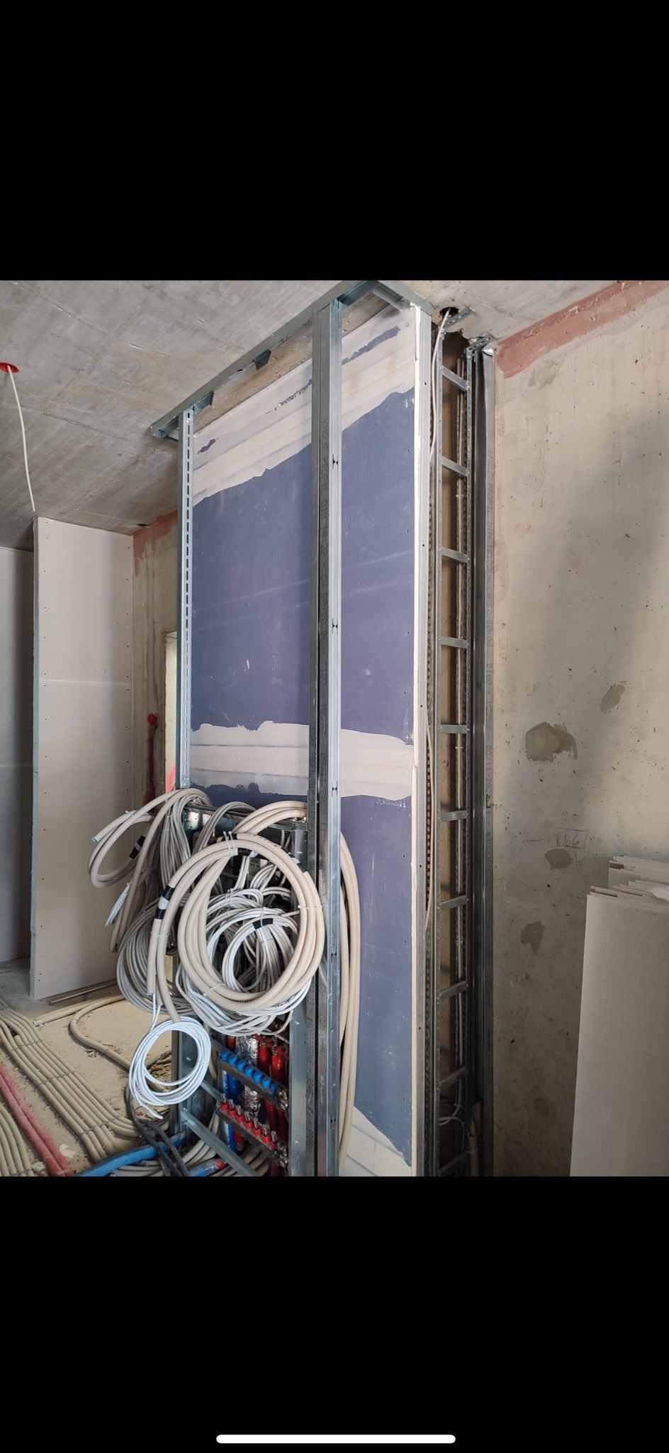Wewnętrzna ściana z płyt gipsowo-kartonowych w trakcie montażu, widoczne metalowe profile, izolacja, instalacje elektryczne i hydrauliczne w wiązkach kabli i rur.