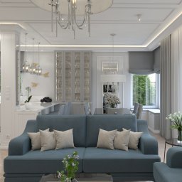 TK design Tomasz Kurpas - Eleganckie wnętrze mieszkania w odcieniach szarości i błękitu z nowoczesnym oświetleniem, wygodną sofą z poduszkami i widokiem na kuchnię z wyspą.