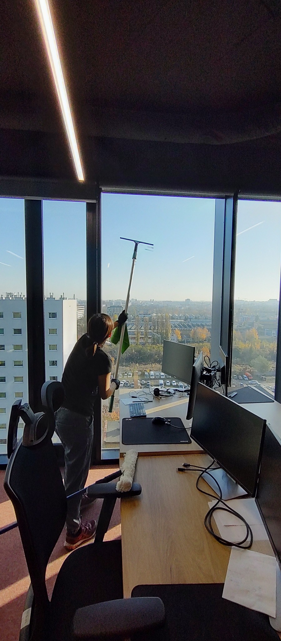 Pracownik w rękawicach czyści okno w biurze za pomocą ściągaczki z długim uchwytem, widok na panoramę miasta za oknem.