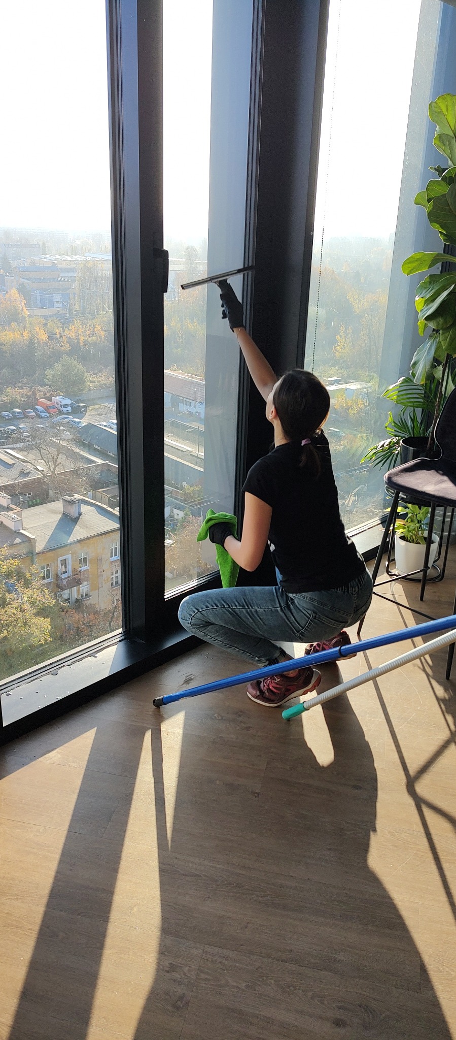 Kobieta w czarnej koszulce i jeansach, w czarnych rękawicach, myje okno w biurze za pomocą ściągaczki i zielonej ścierki. Widok na miasto przez okno.