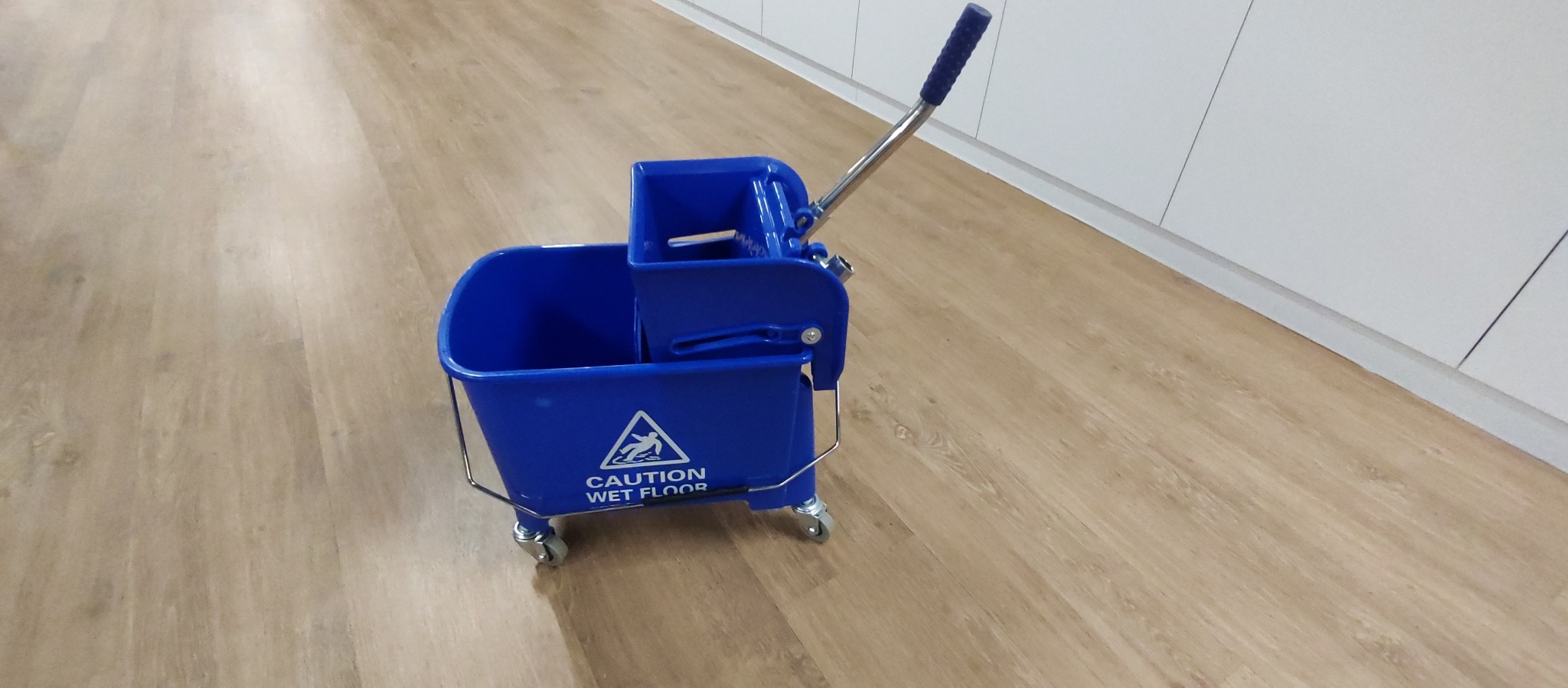 Niebieski wózek do mycia podłóg z wyciskarką, stojący na jasnej podłodze drewnopodobnej w biurze, widoczny znak ostrzegawczy 'Caution Wet Floor'.