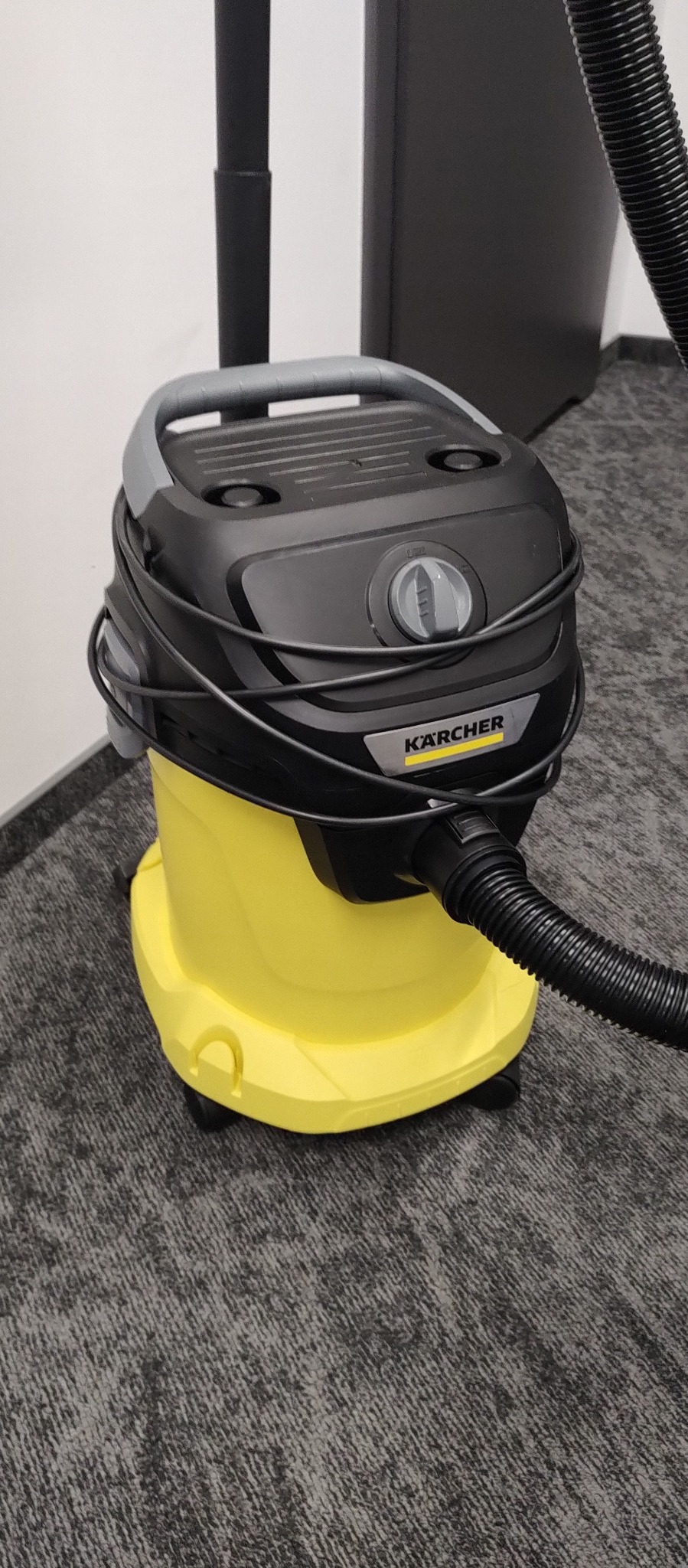 Żółto-czarny odkurzacz Kärcher z owiniętym wężem, stojący na szarym dywanie w biurze.