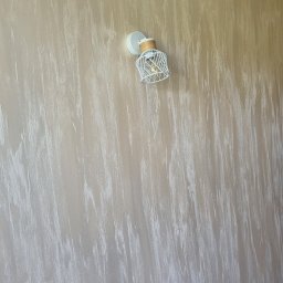 Przemysław winiarowski ogólnobudowlana - Beżowa ściana z dekoracyjną strukturą przypominającą przecierkę, oświetlona nowoczesnym kinkietem z metalowym ażurem w kształcie klatki.