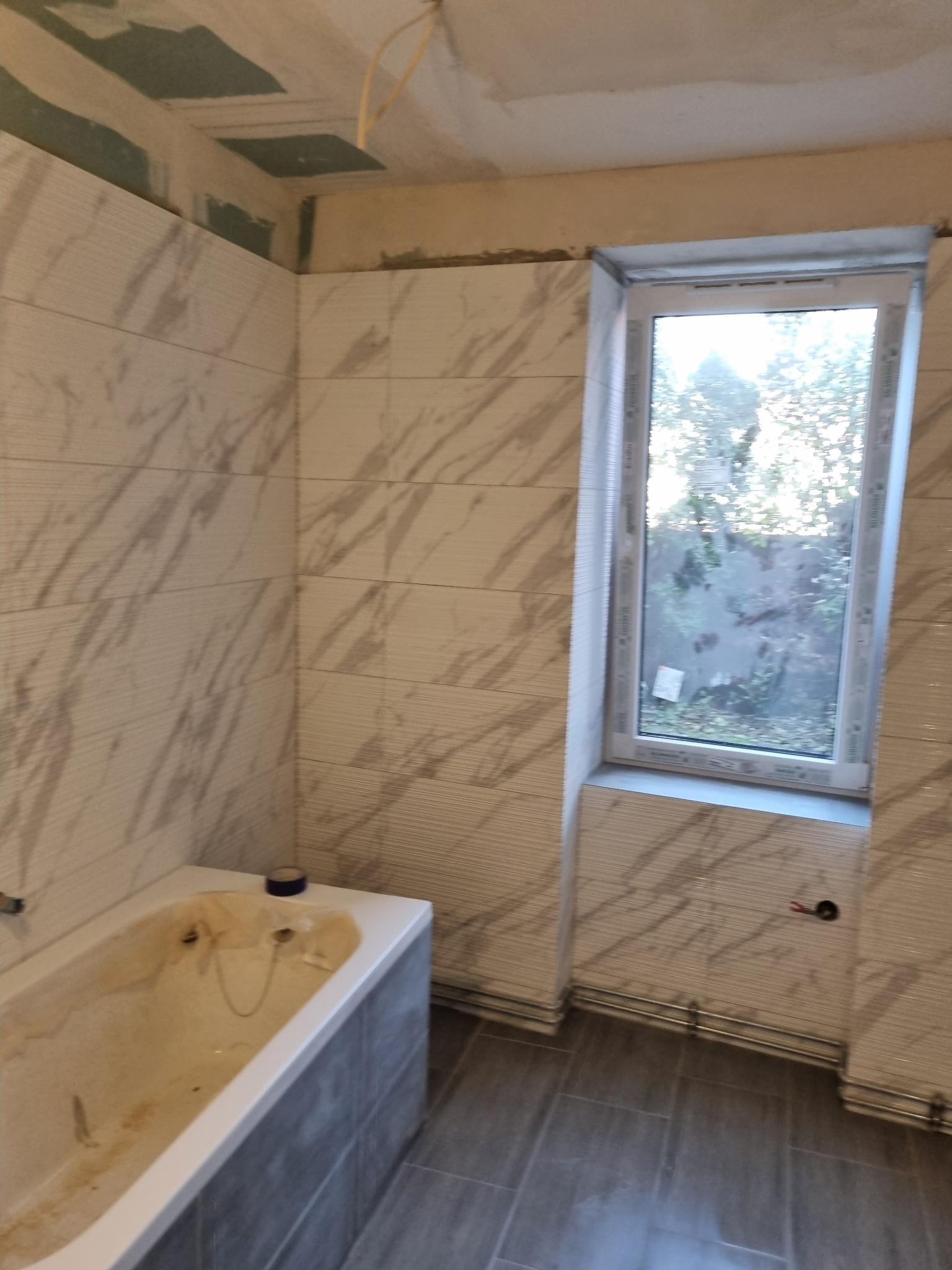 Niedokończona łazienka z wanną, marmurkowymi płytkami na ścianach i szarymi płytkami podłogowymi, widoczne niedociągnięcia na suficie i wystające przewody elektryczne.