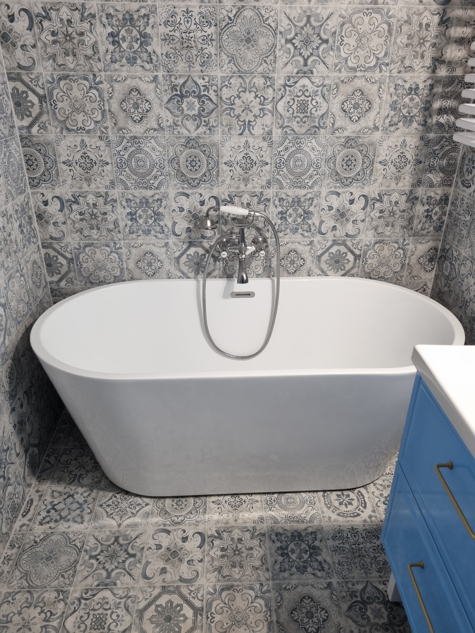 Wolnostojąca wanna z chromowaną baterią na tle ściany i podłogi wyłożonej dekoracyjnymi, wzorzystymi płytkami w odcieniach szarości i błękitu; widoczny fragment niebieskiej szafki z złotymi uchwytami.