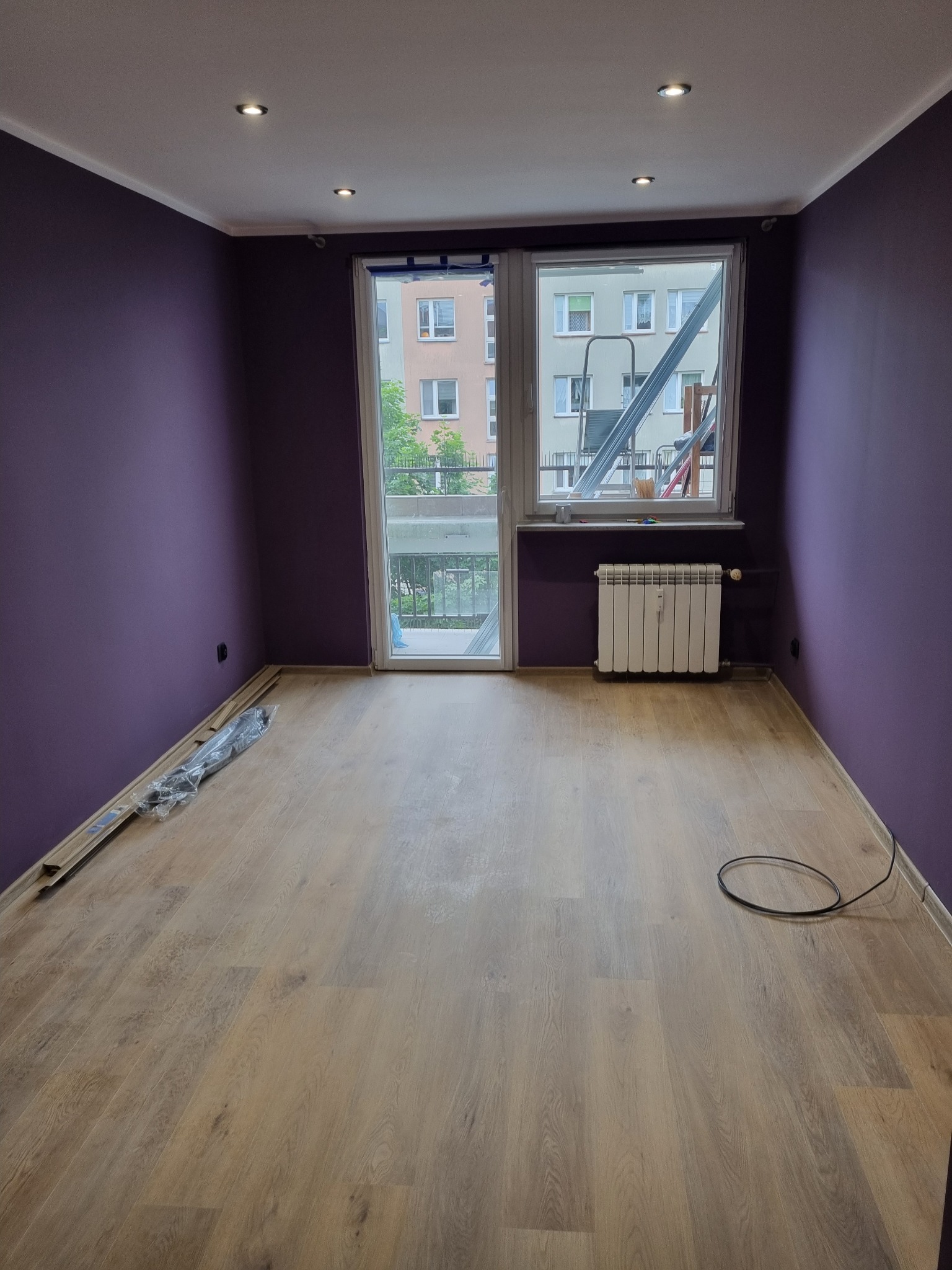 Pusty pokój po remoncie z fioletowymi ścianami, panelami podłogowymi w odcieniach brązu i beżu, grzejnikiem pod oknem balkonowym i widocznymi listwami przypodłogowymi w opakowaniu.