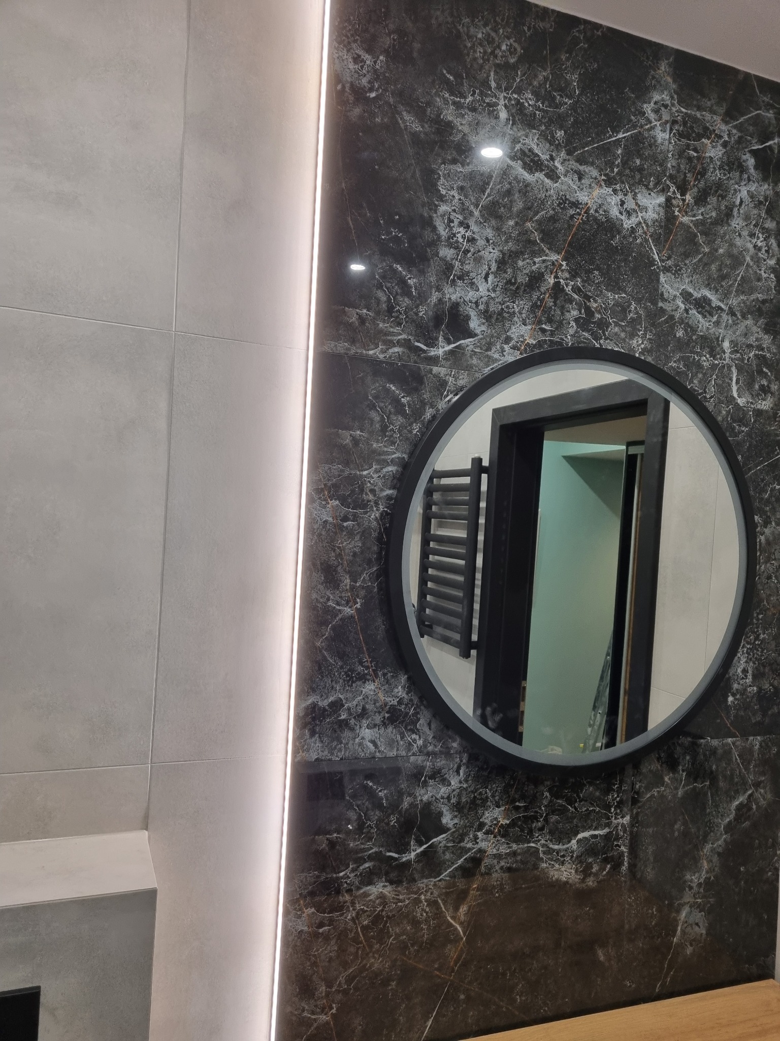 Nowoczesna łazienka z szarymi i czarnymi płytkami imitującymi marmur, oświetleniem LED w pionowej szczelinie, okrągłym lustrem z widocznym grzejnikiem drabinkowym i fragmentem blatu.