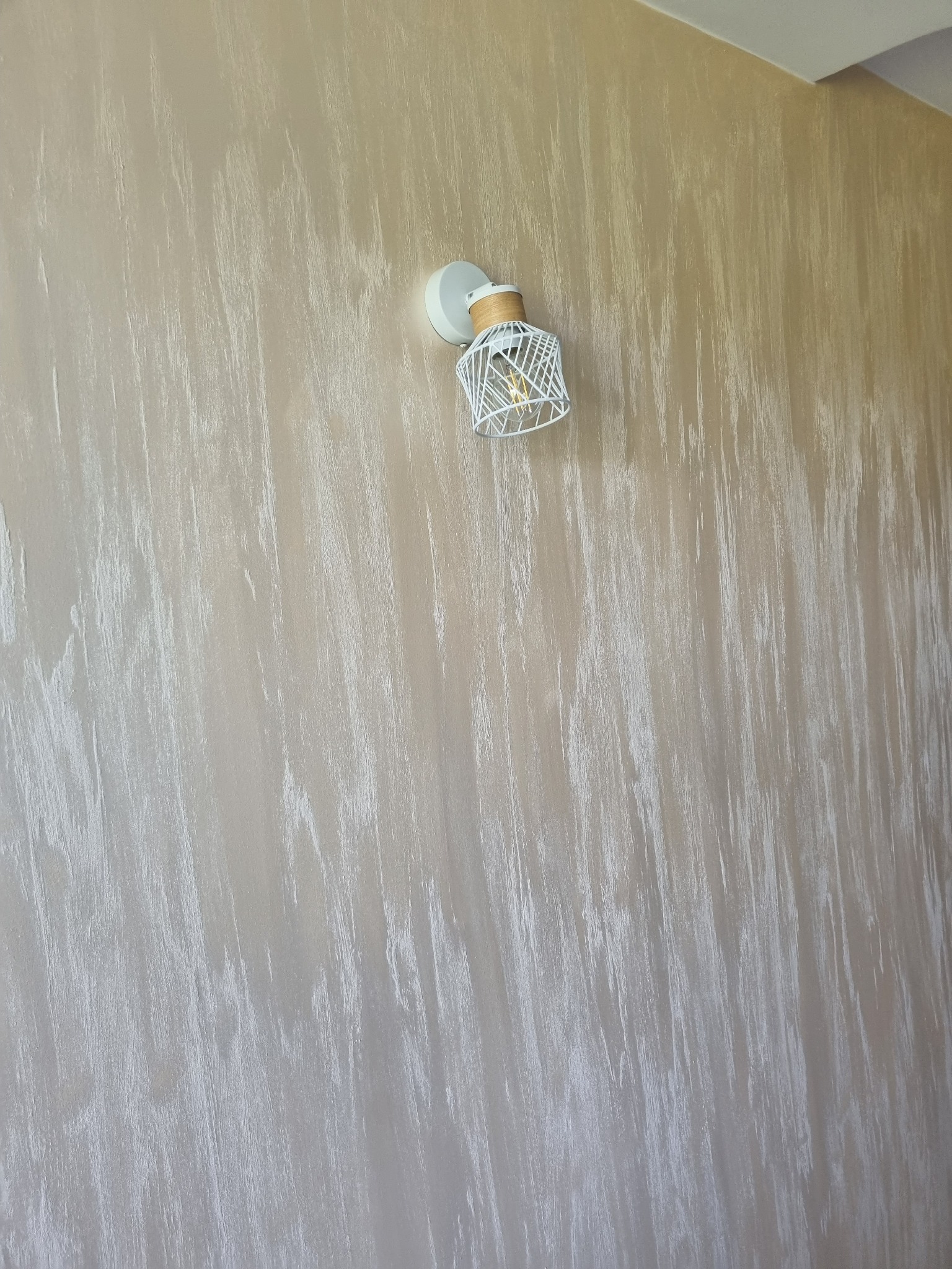 Beżowa ściana z dekoracyjną strukturą przypominającą przecierkę, oświetlona nowoczesnym kinkietem z metalowym ażurem w kształcie klatki.